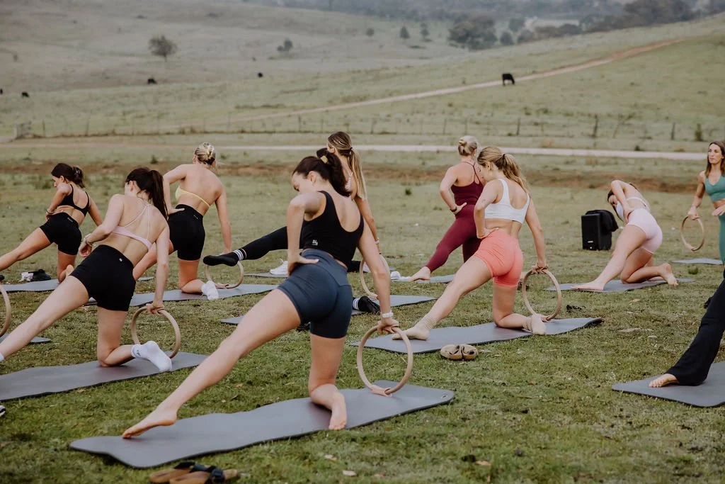 Women participating in an outdoor yoga class at Mudgee Glenayr retreat NSW, using yoga rings, with rolling hills and grazing cows in the background.