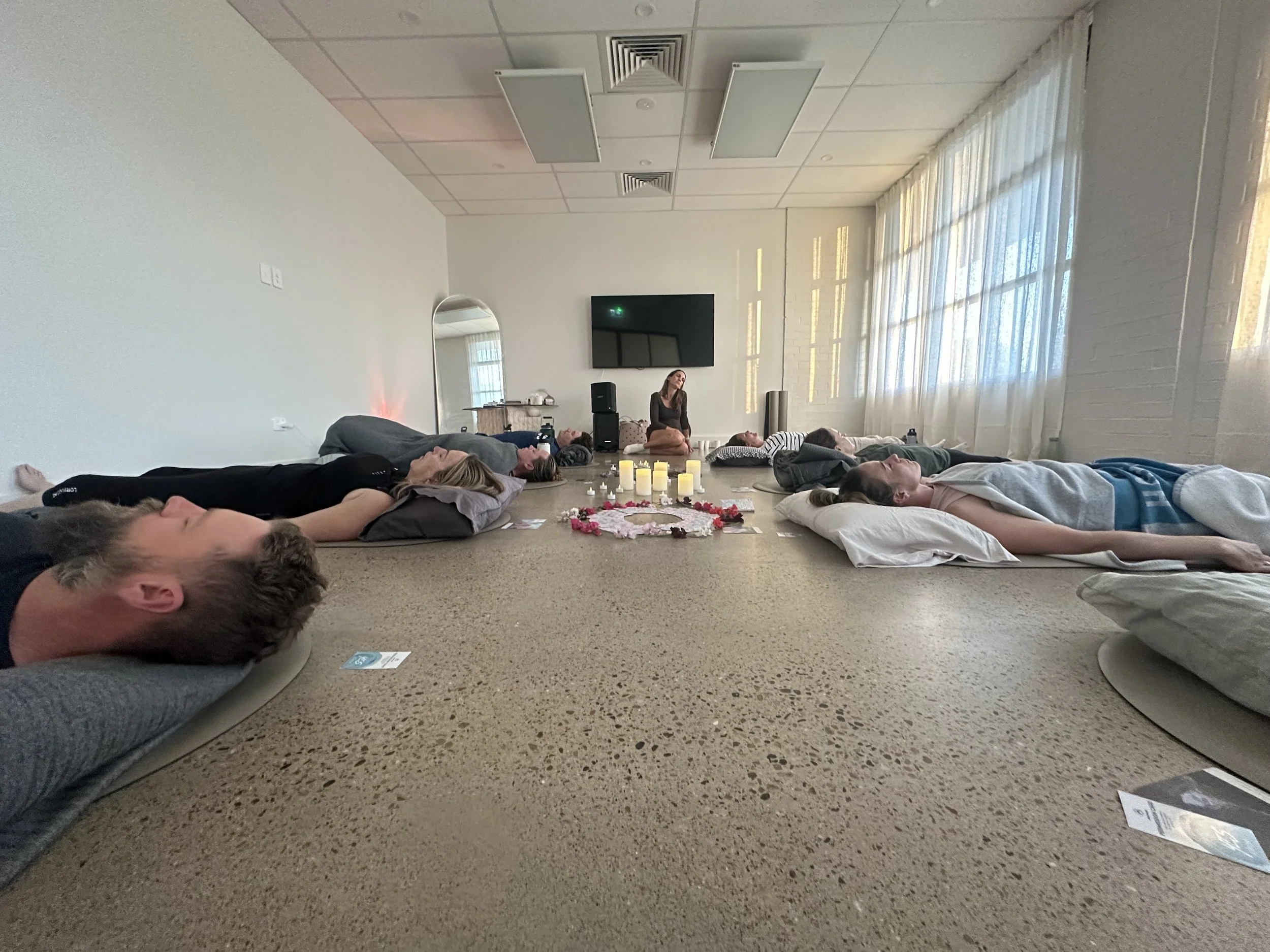 A group of people lying on their backs on yoga mats in a spacious room with white walls and large windows, participating in a meditation or relaxation session. A woman, likely the instructor, is sitting cross-legged at the front near a display of candles and flowers.