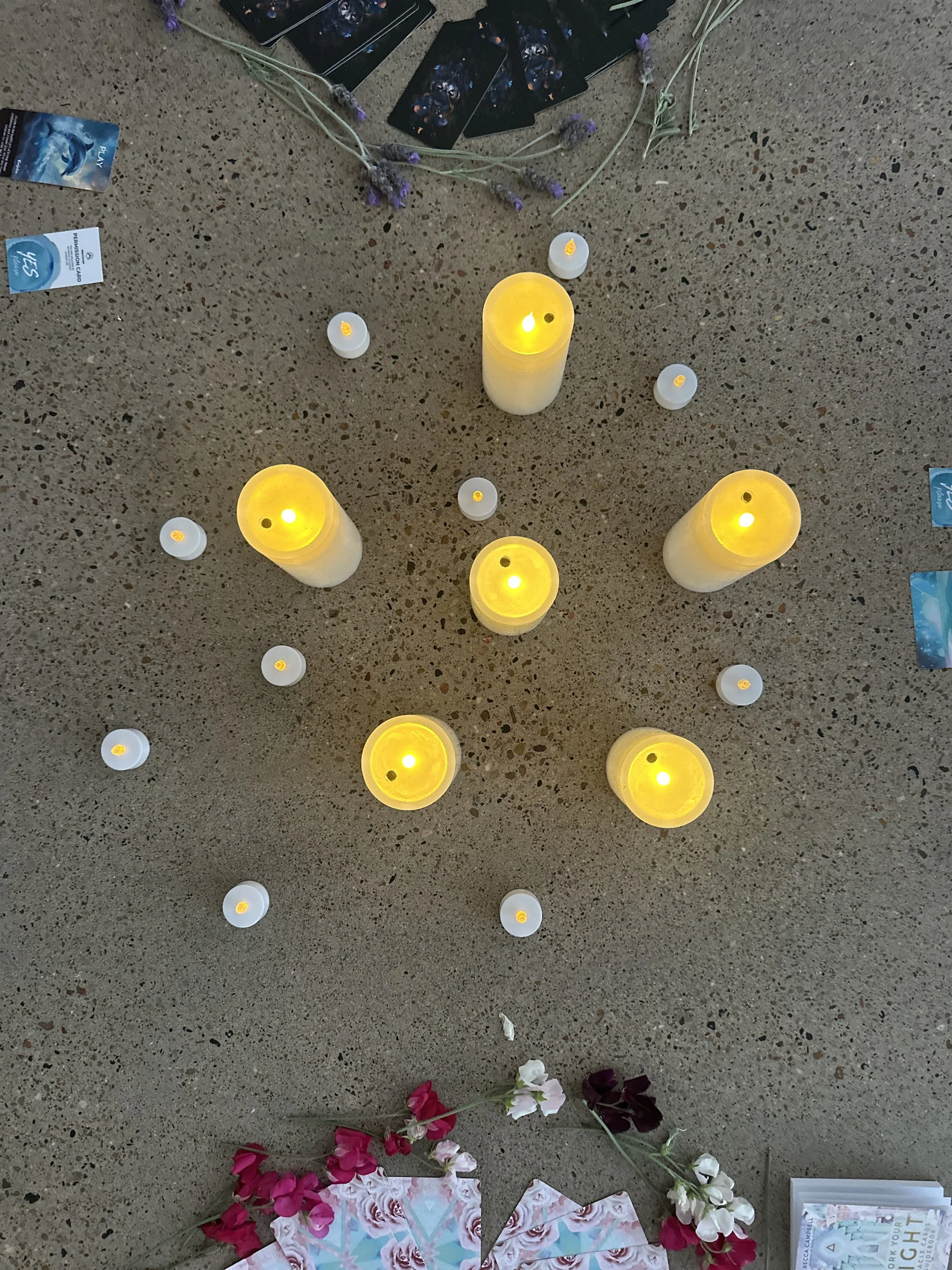 Candlelight vigil with seven lit yellow candles arranged in a cross pattern on a concrete surface. Surrounding the candles are small white tealight candles, tarot cards, flowers, and a few scattered cards and papers.