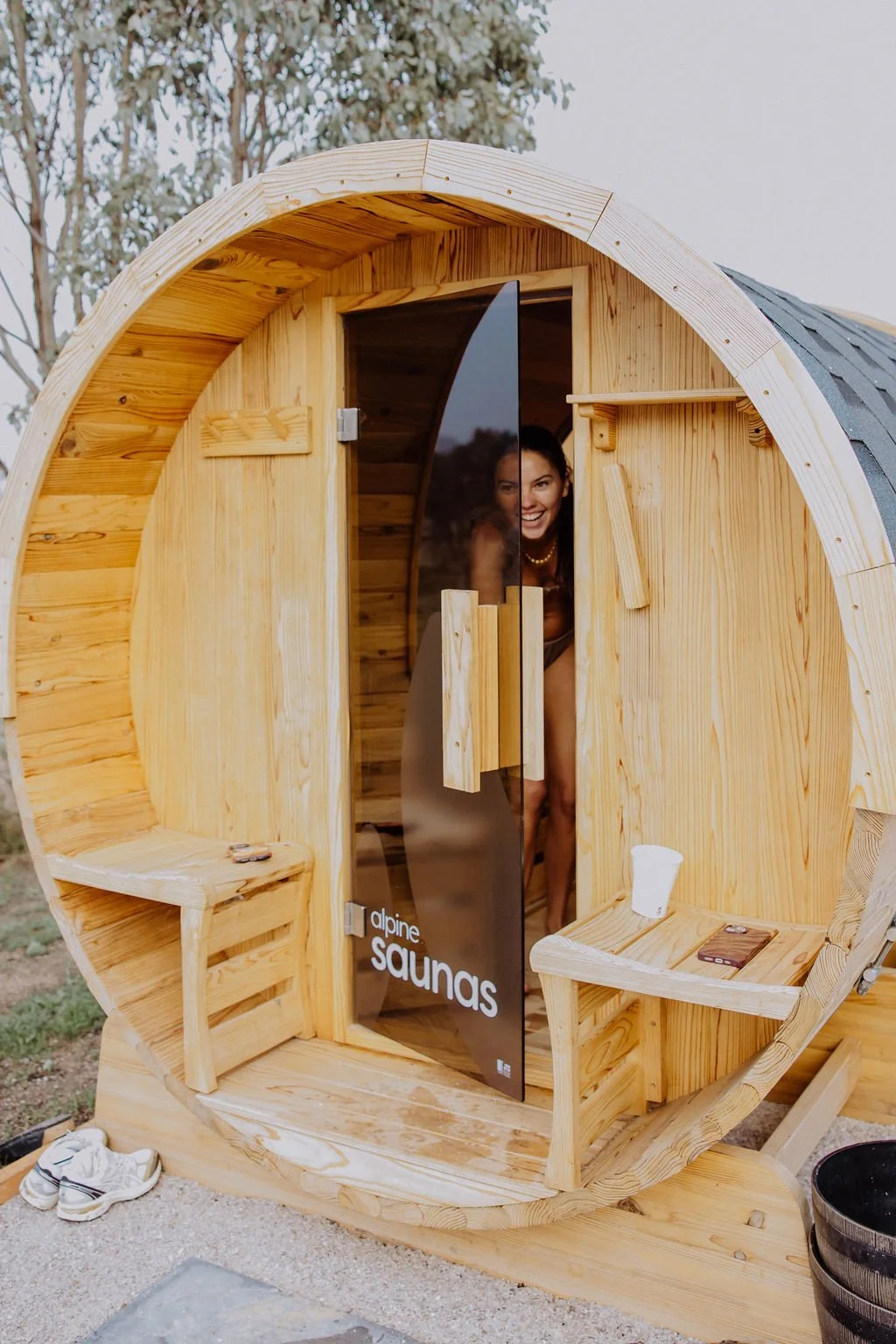A woman smiling inside a small wooden sauna at a wellness retreat in Mudgee with a glass door, outdoors, with trees in the background.