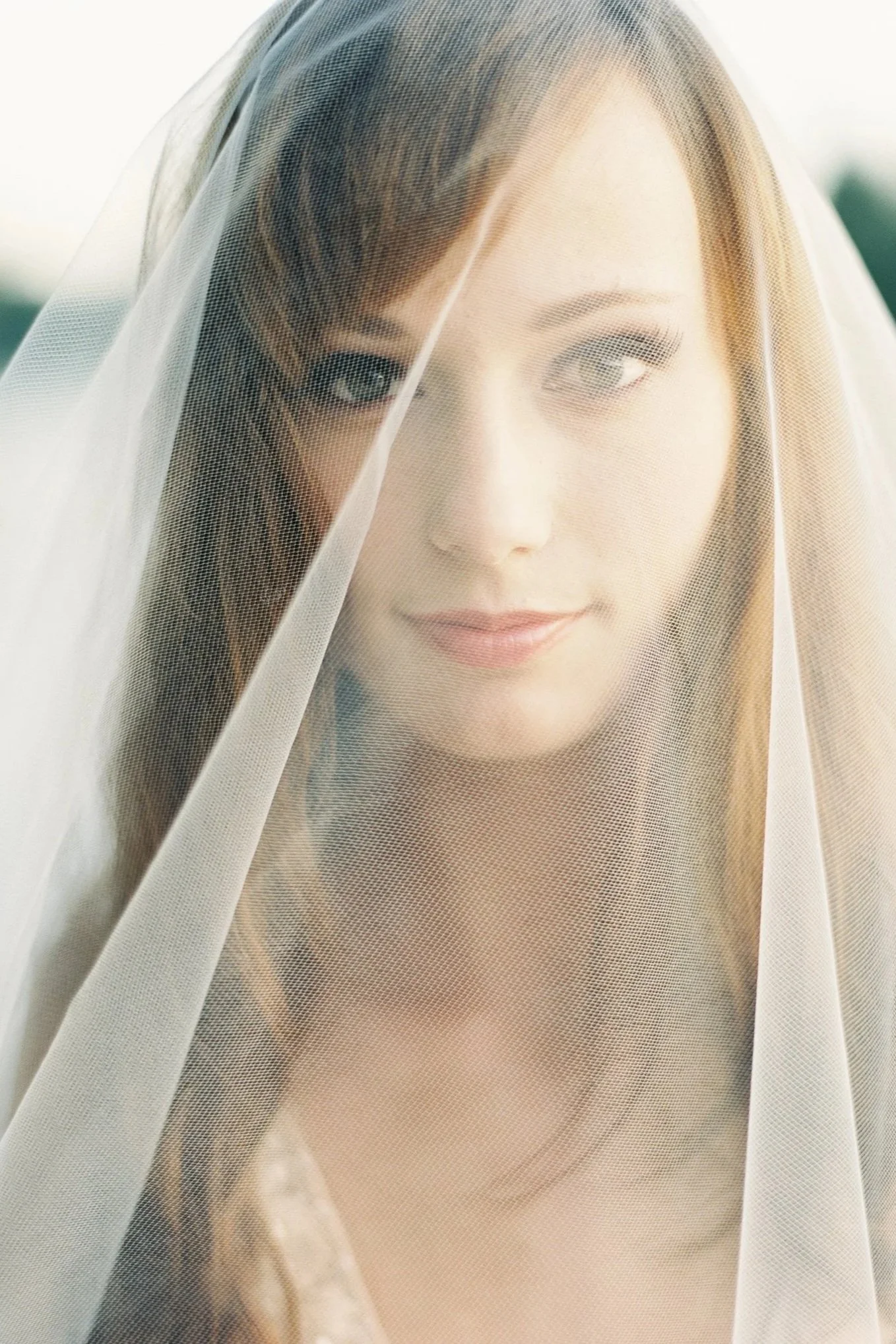 A woman with red hair and light blue eyes, wearing a sheer veil over her face, smiling gently outdoors.