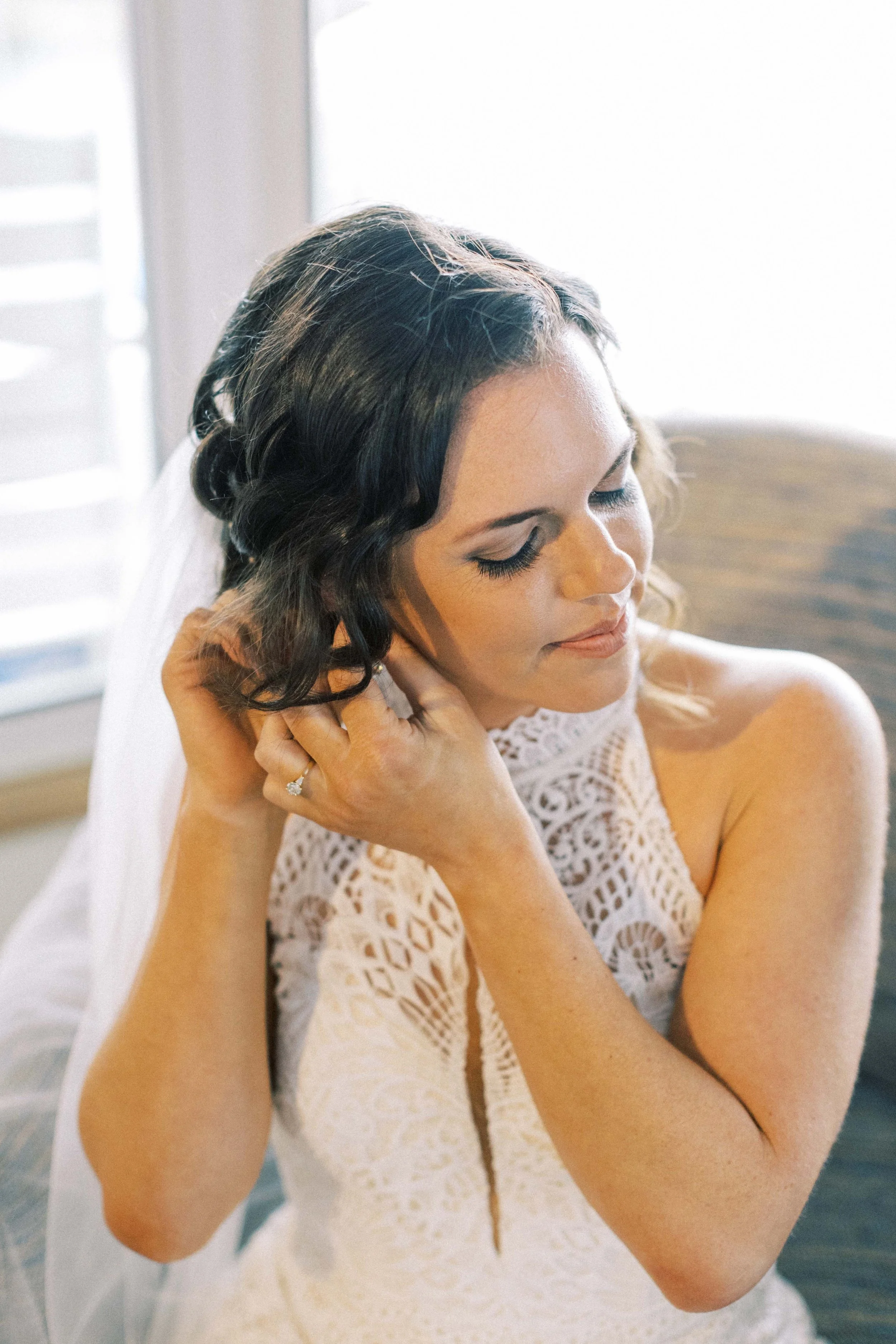 Bride with soft curls touching her earring, wearing natural makeup and an elegant sleeveless gown by Spokane and Coeur d’Alene bridal hair and makeup artist Terri Reece Studios.