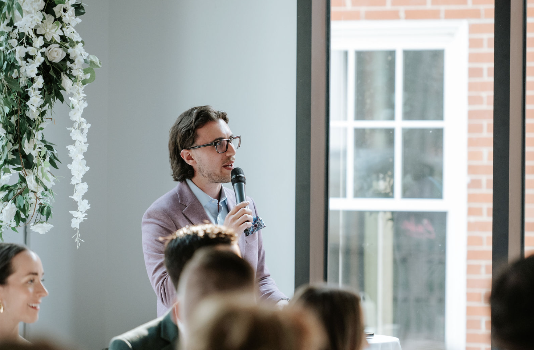 Man with glasses holding a microphone and speaking at an indoor event, with people seated in front of him and a large window behind.