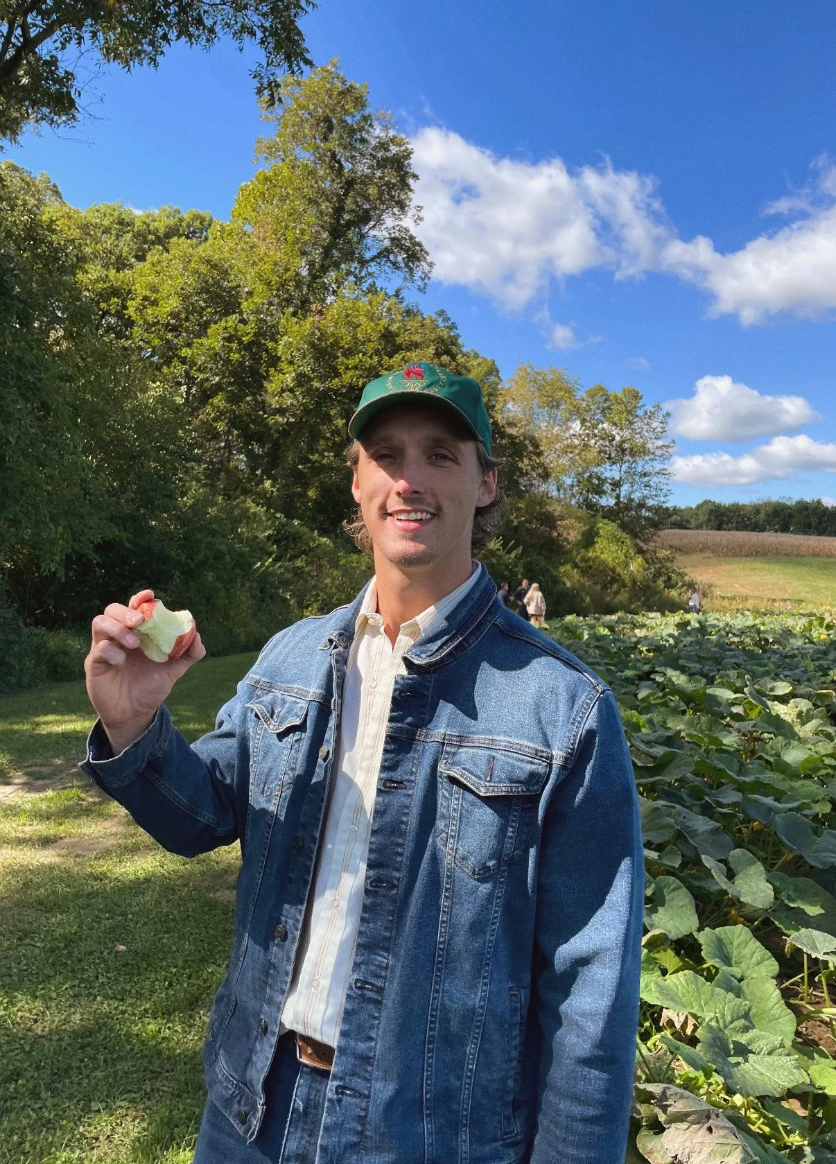 A young man outdoors on a sunny day, smiling and holding an apple core. He is wearing a green cap, a denim jacket, and a light-colored shirt, with a field of green plants and trees in the background.