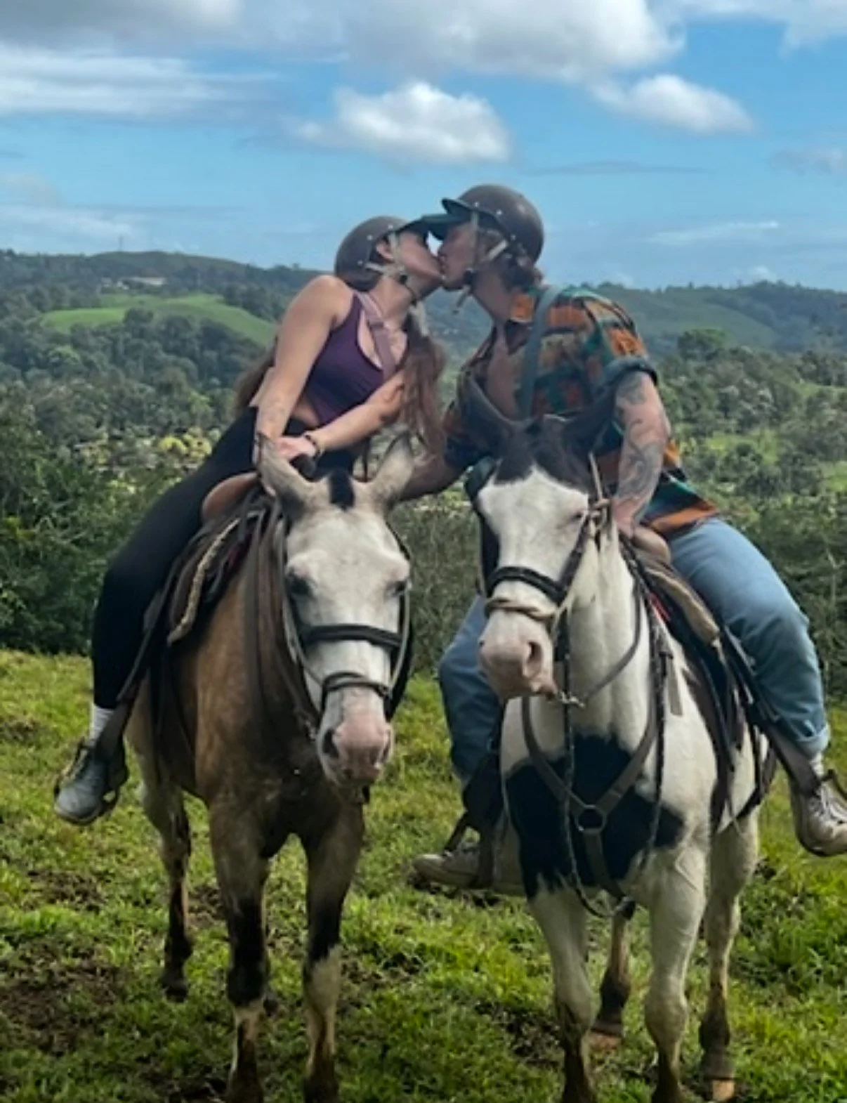 A couple on horseback kissing with a scenic green landscape and blue sky in the background.