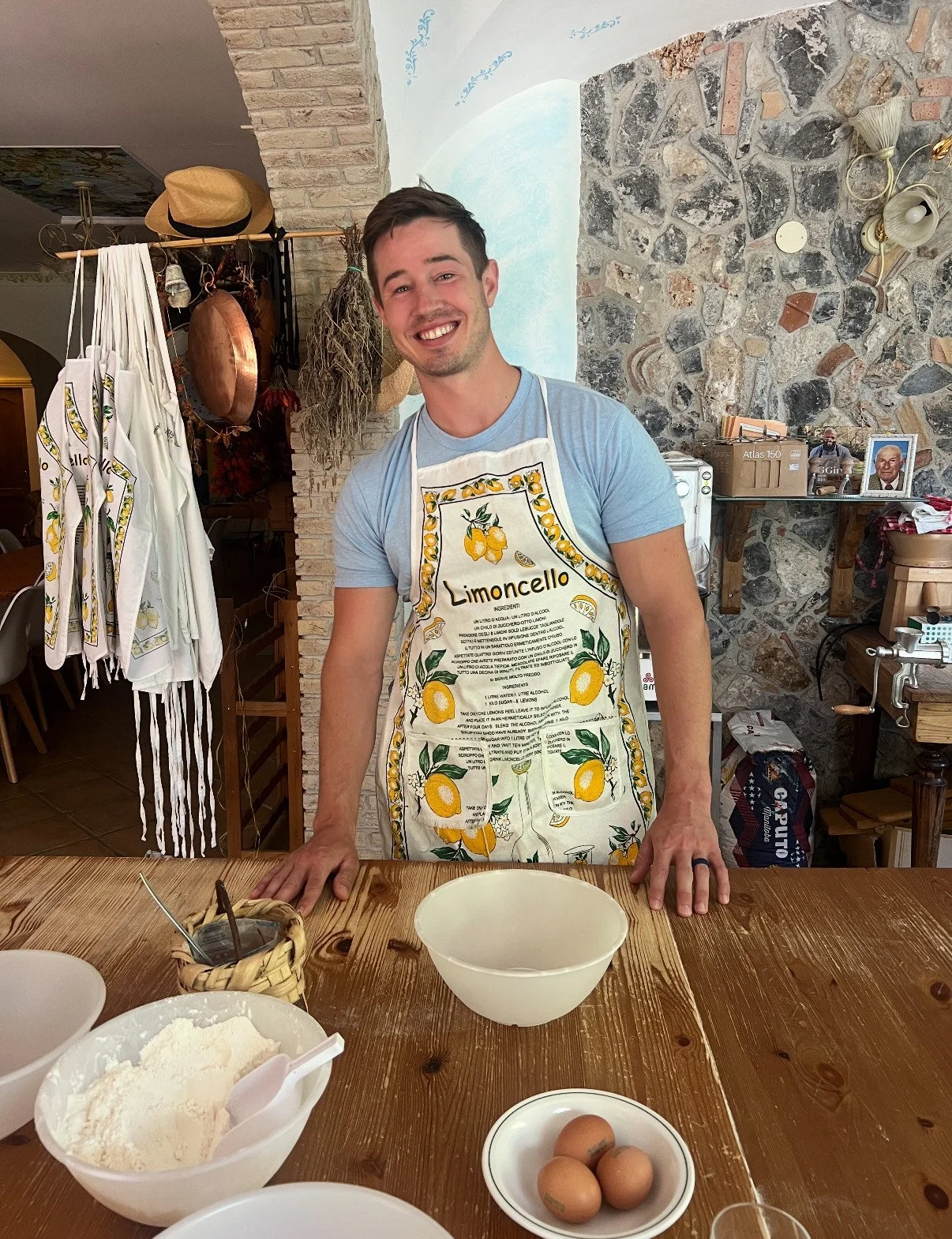 A smiling man in a lemon-themed apron standing at a wooden table with baking ingredients and eggs, inside a cozy kitchen.