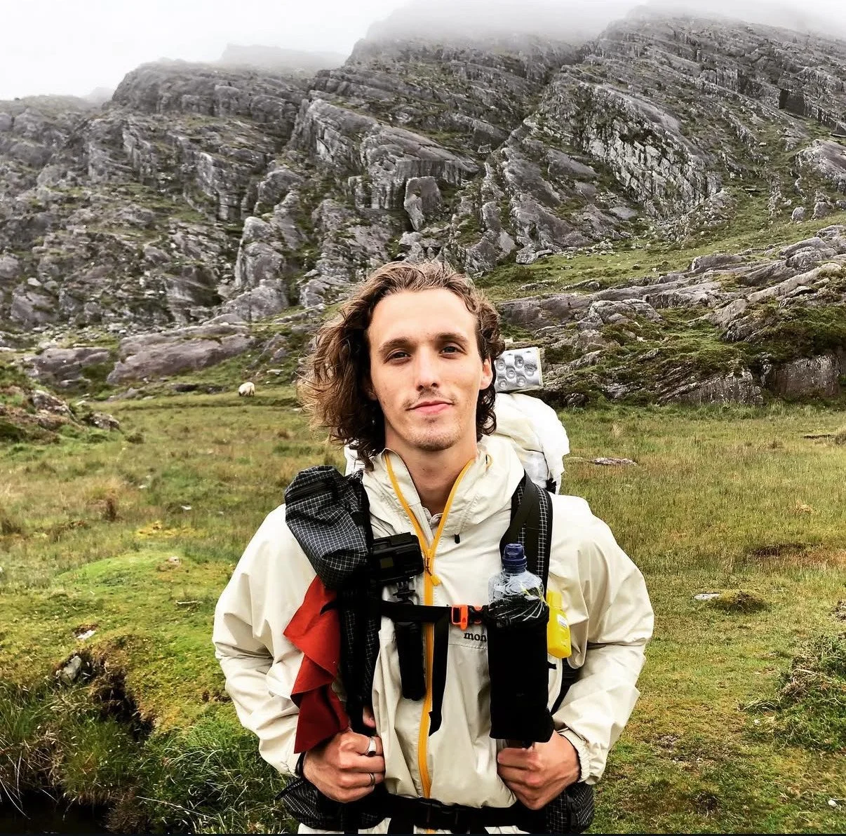 A young man with long curly hair and a slight smile standing outdoors in front of a rocky mountain landscape with lush green grass and misty skies.