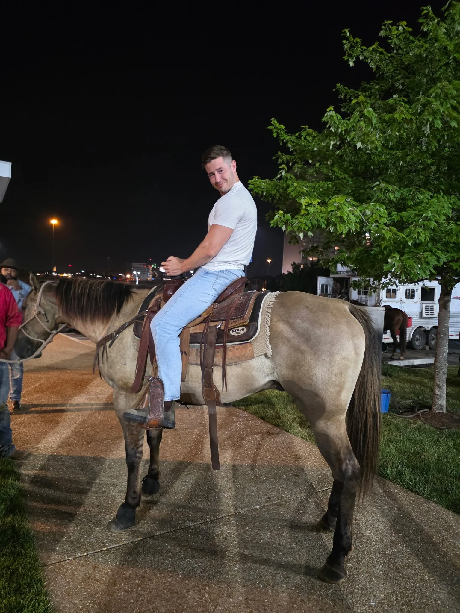A young man in a white t-shirt and light jeans sitting on a beige horse with a brown saddle at night. There is a tree and some vehicles in the background, with city lights and dark sky overhead.