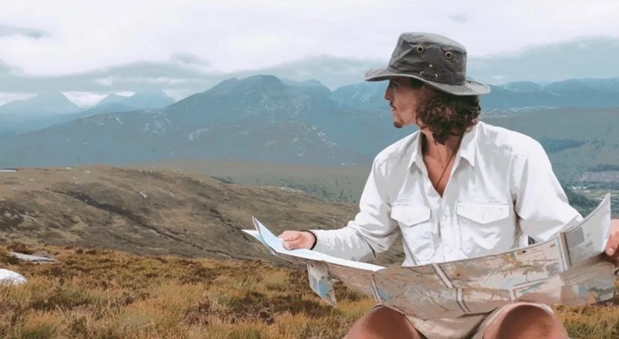 A man in a white shirt and wide-brimmed hat sitting outdoors, studying a large map with mountains in the background.