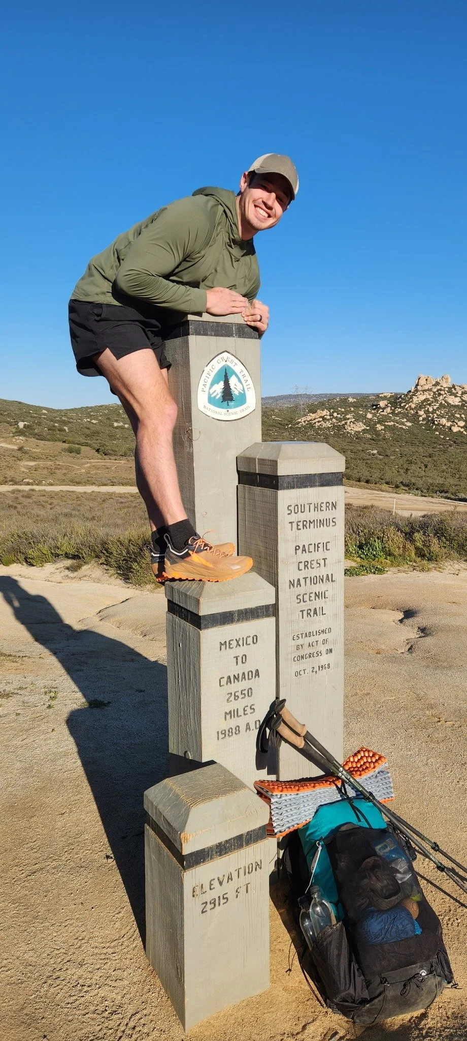 Hiker standing on a trail marker at the southern terminus of the Pacific Crest National Scenic Trail, near the Mexico-Canada border, with a backpack and hiking poles, in a scenic landscape under a clear blue sky.