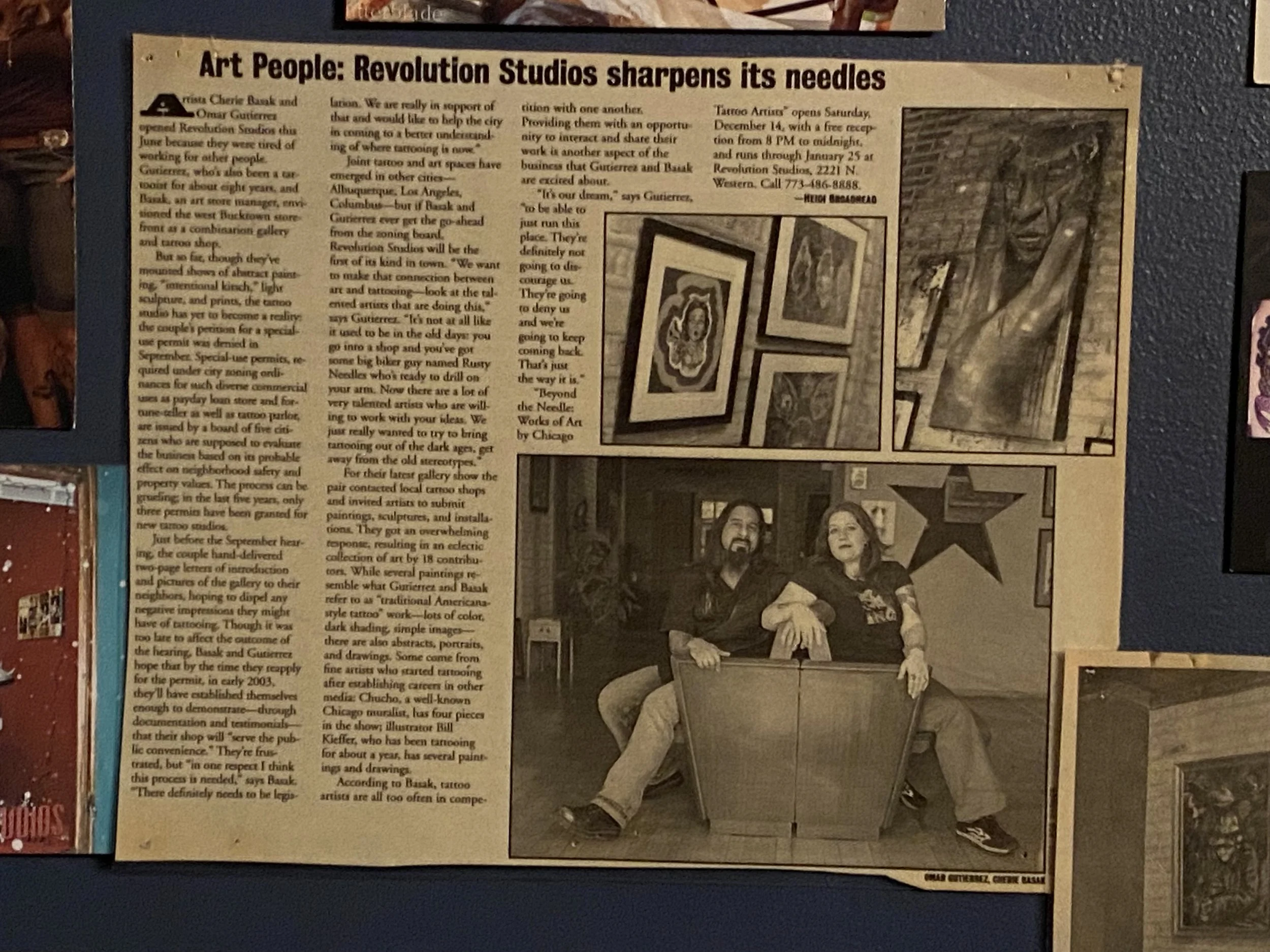 A newspaper article titled "Art People: Revolution Studios sharpens its needles" with text and four framed art pieces and a photo of a man and woman sitting on a bench. The background features a brick wall and other artworks.