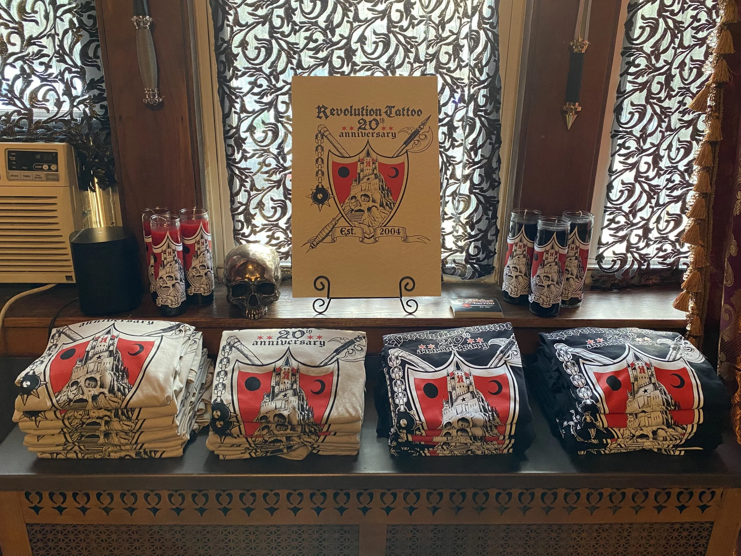 Display of merchandise celebrating the 20th anniversary of Revolution Tattoo, including T-shirts, cups, and a poster, with decorative curtains in the background.