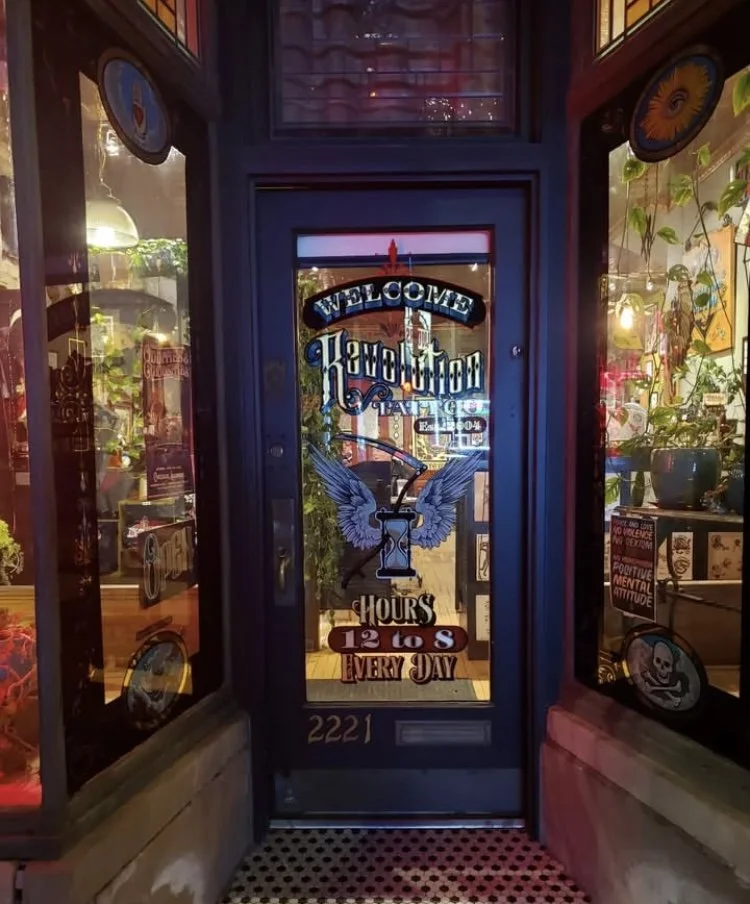 Front door of a tattoo shop with colorful graphics and signs, including a welcome message, store hours, and decorative stickers like a skull and angel wings.
