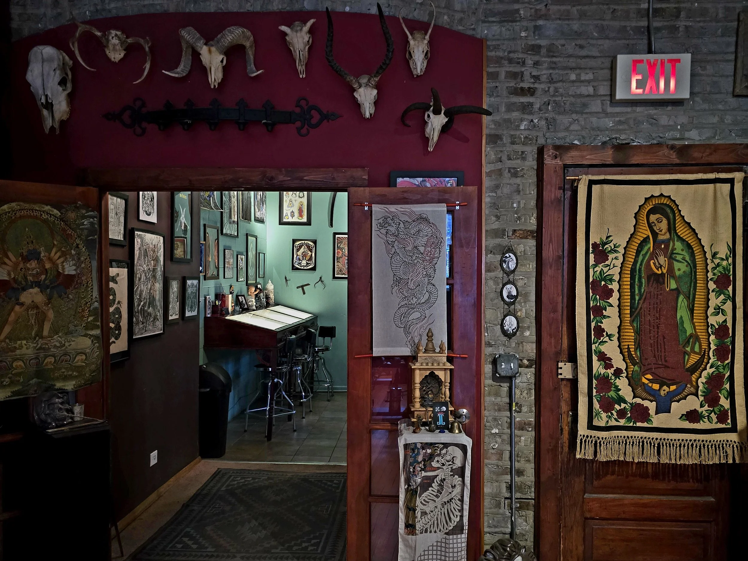 Interior of a tattoo parlor with tattoo artwork on the walls, mounted animal skulls on the wall, a tapestry of Our Lady of Guadalupe, and a small altar with religious figures and decorations.
