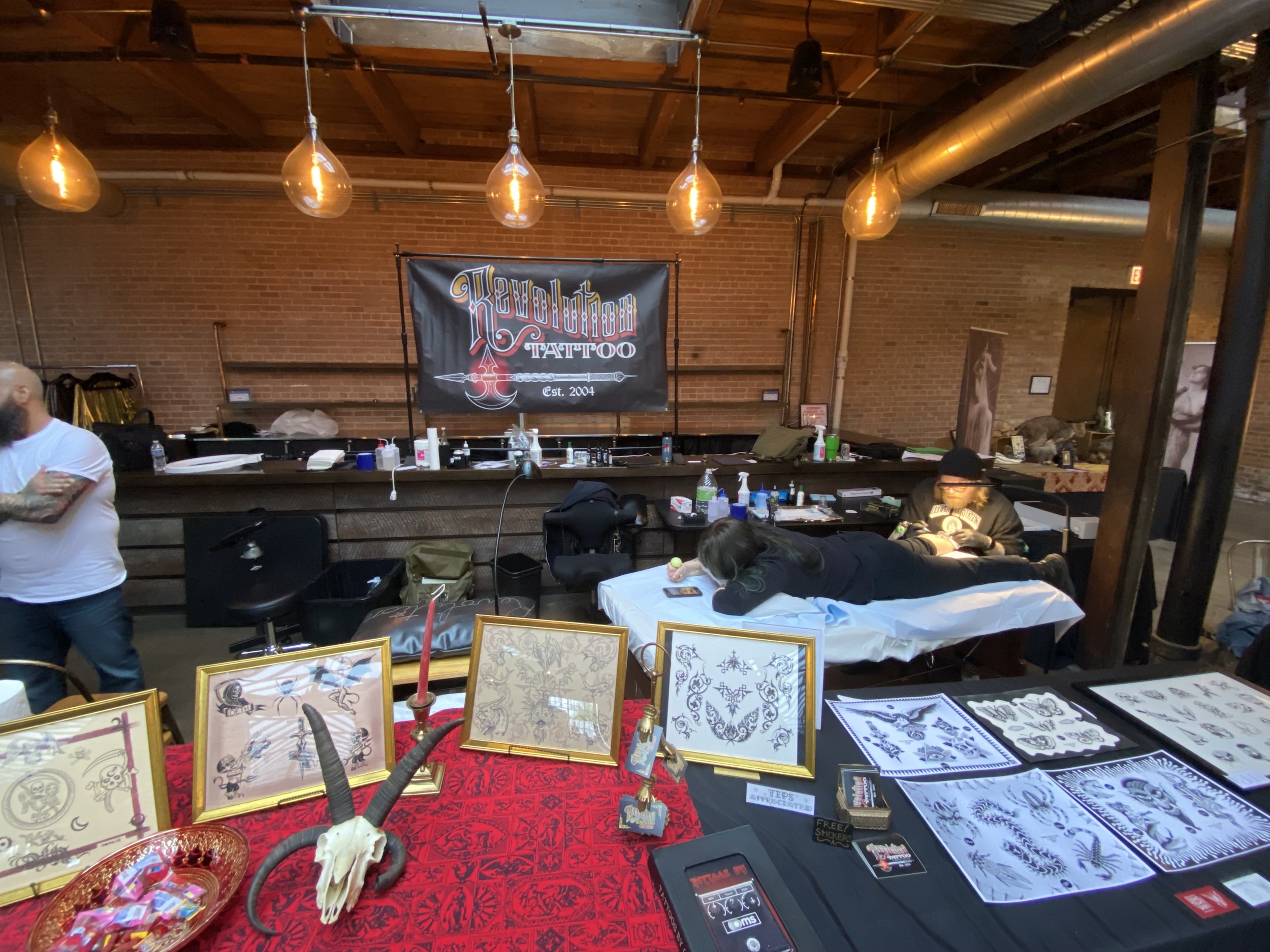 Tattoo studio interior with framed tattoo art on display, a black banner reading "Revolution Tattoo Est. 2004" hanging on a brick wall, tattoo artists working on clients, and tattoo supplies on a counter.