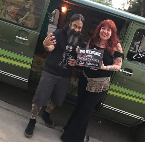 A man and woman standing outside next to a green bus, holding a sign that reads "The Woodlands 2019 People's Choice". The man has long hair, a beard, tattoos, and is making a peace sign. The woman has red hair and is smiling.