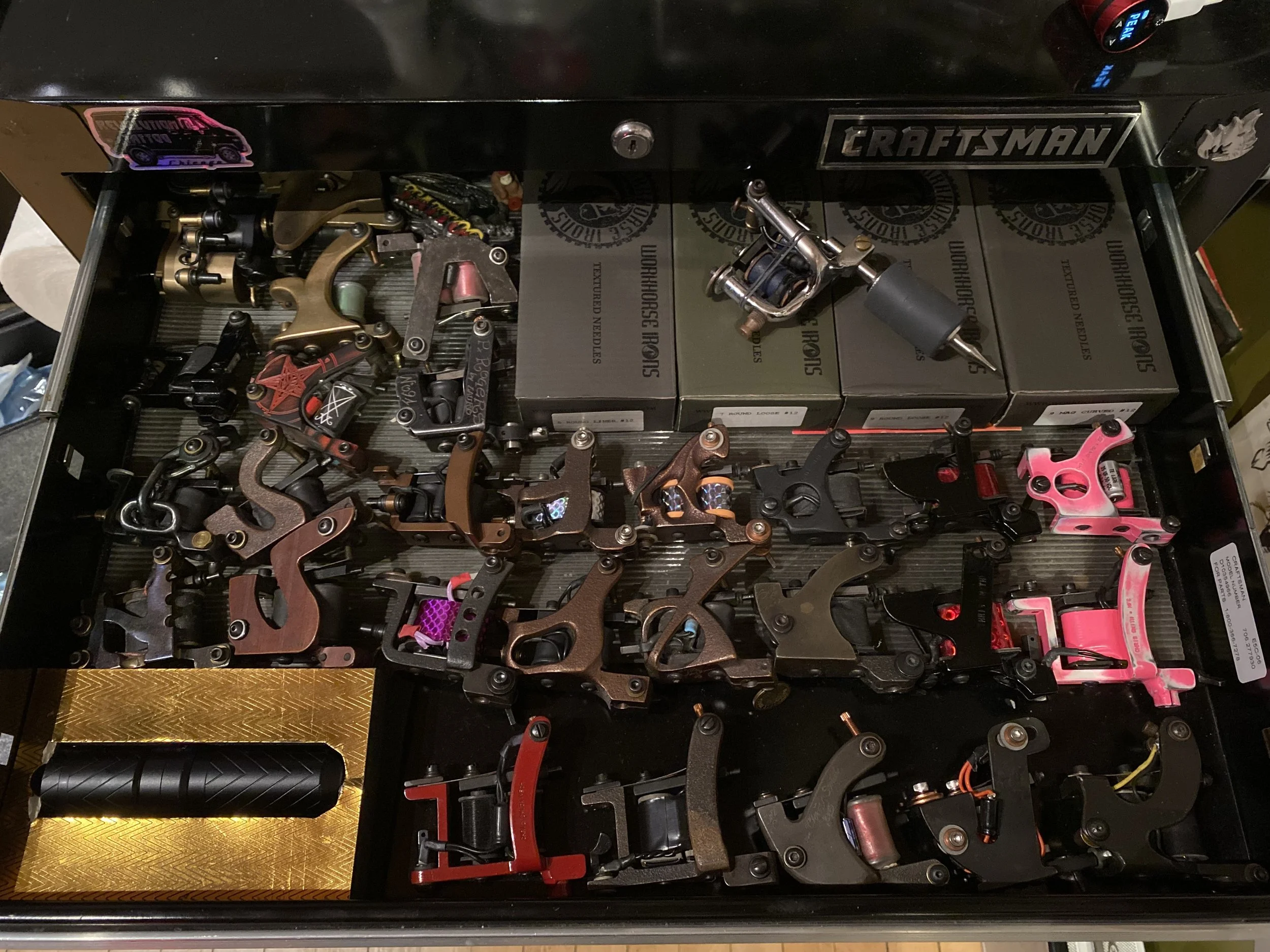 A drawer containing many parts for tattoo machines, including frames, coils, and other components, organized in rows and boxes.