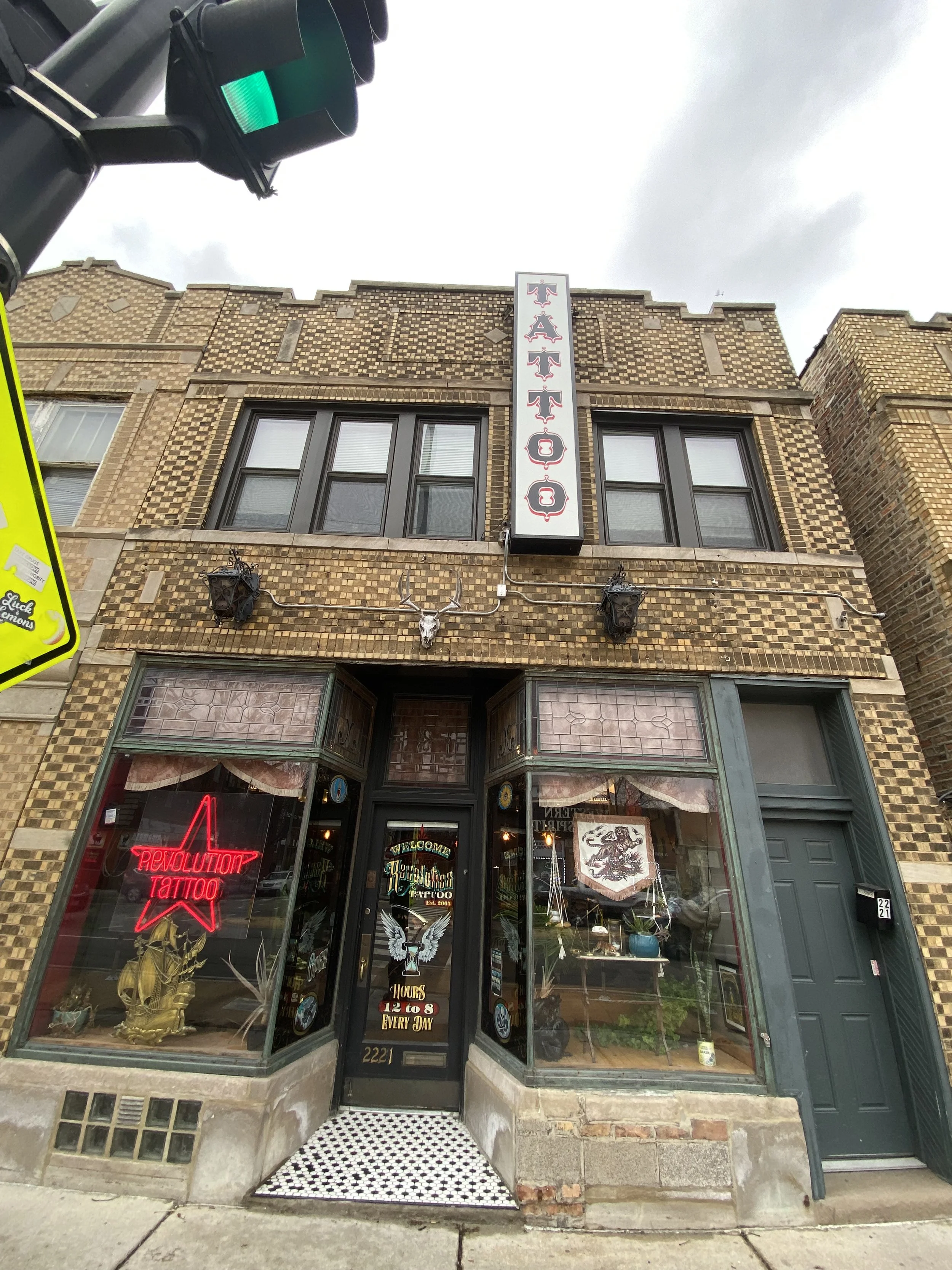 Front of a tattoo studio in a brick building with a vertical sign reading 'TATTOO', two black decorative lanterns, a mounted deer skull with antlers above the entrance, neon sign saying 'REVOLUTION TATTOO', and windows displaying tattoo artwork.