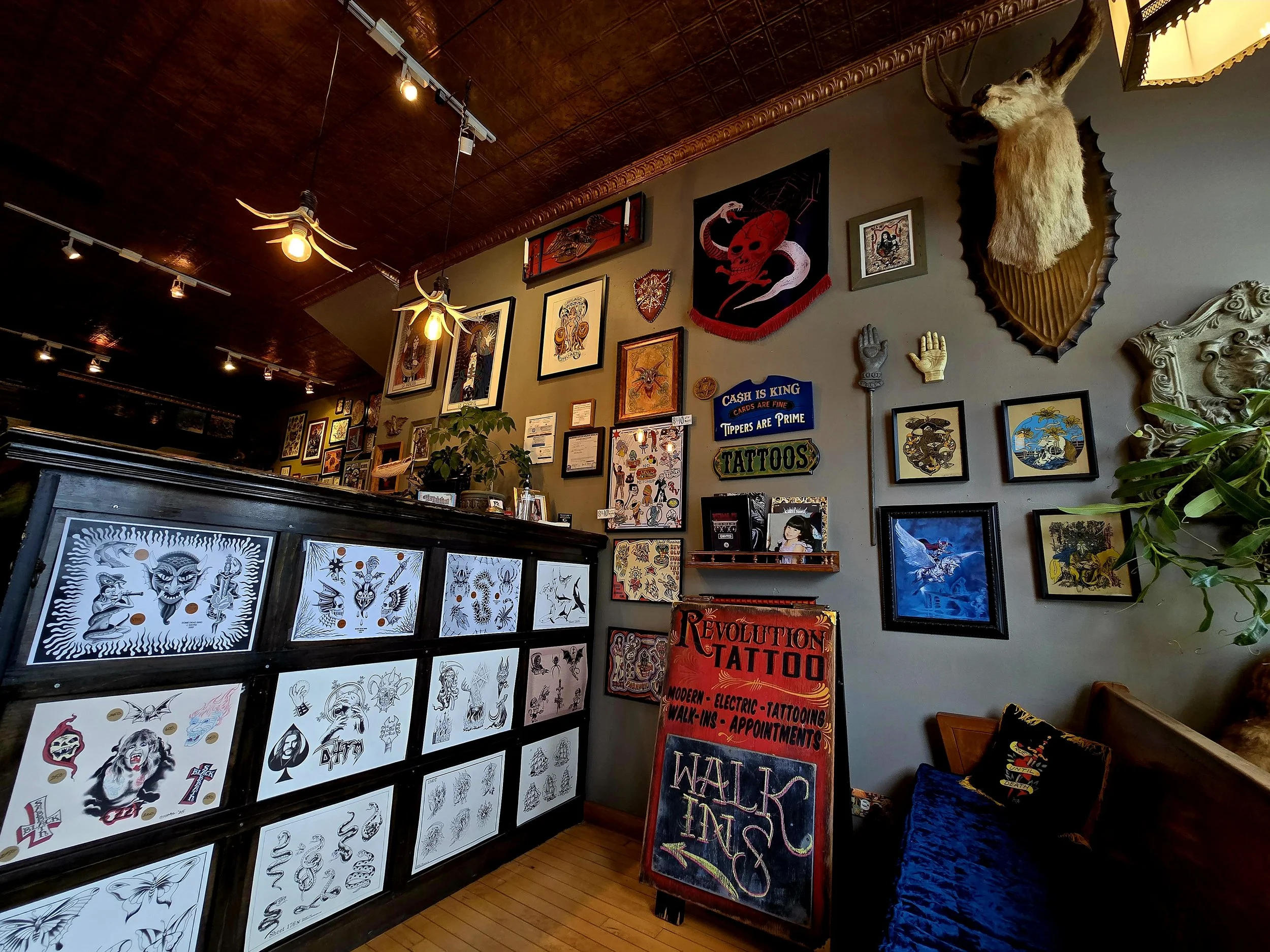 Tattoo studio with framed artwork, animal mounts, and tattoo signs, including a red and black REVOLUTION TATTOO sign, and a chalkboard sign with 'WALK INS' written on it.