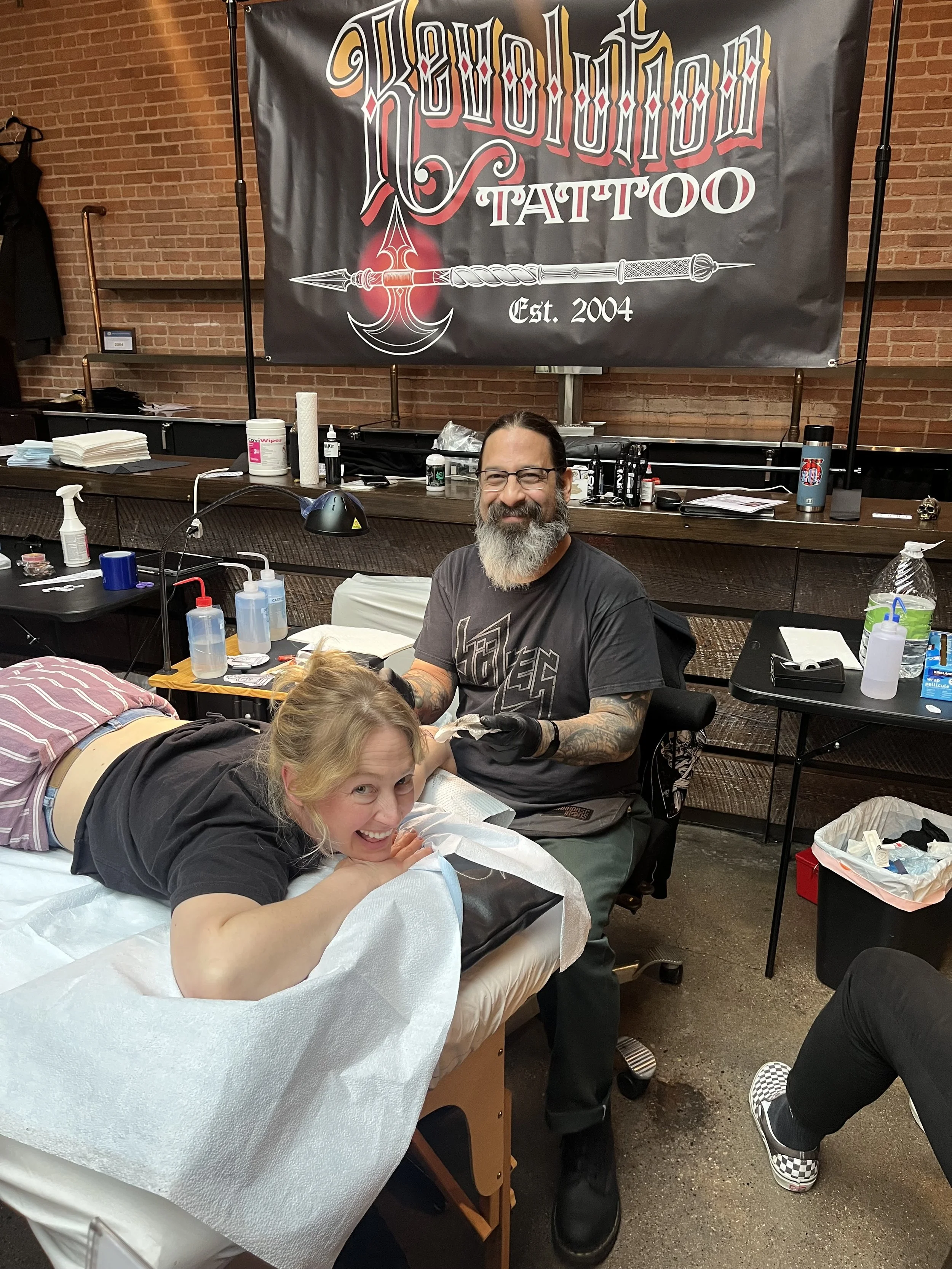 A woman lying on a tattoo bed, smiling, as a tattoo artist with glasses and tattoos works on her arm in a tattoo studio with a "Revolution Tattoo" banner in the background.