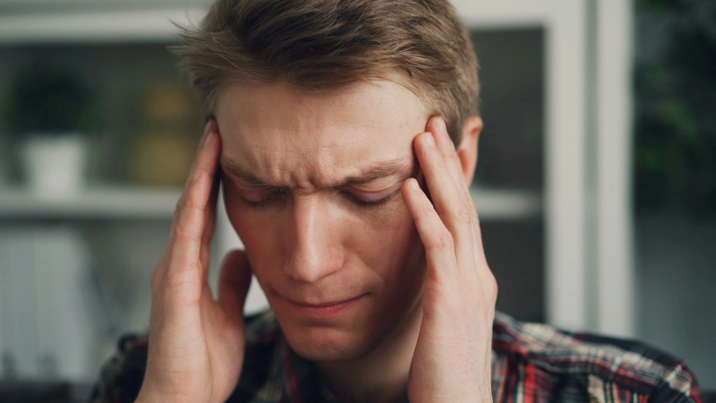 A young man with closed eyes holds his head with both hands, appearing stressed or in pain, indoors with blurred background.