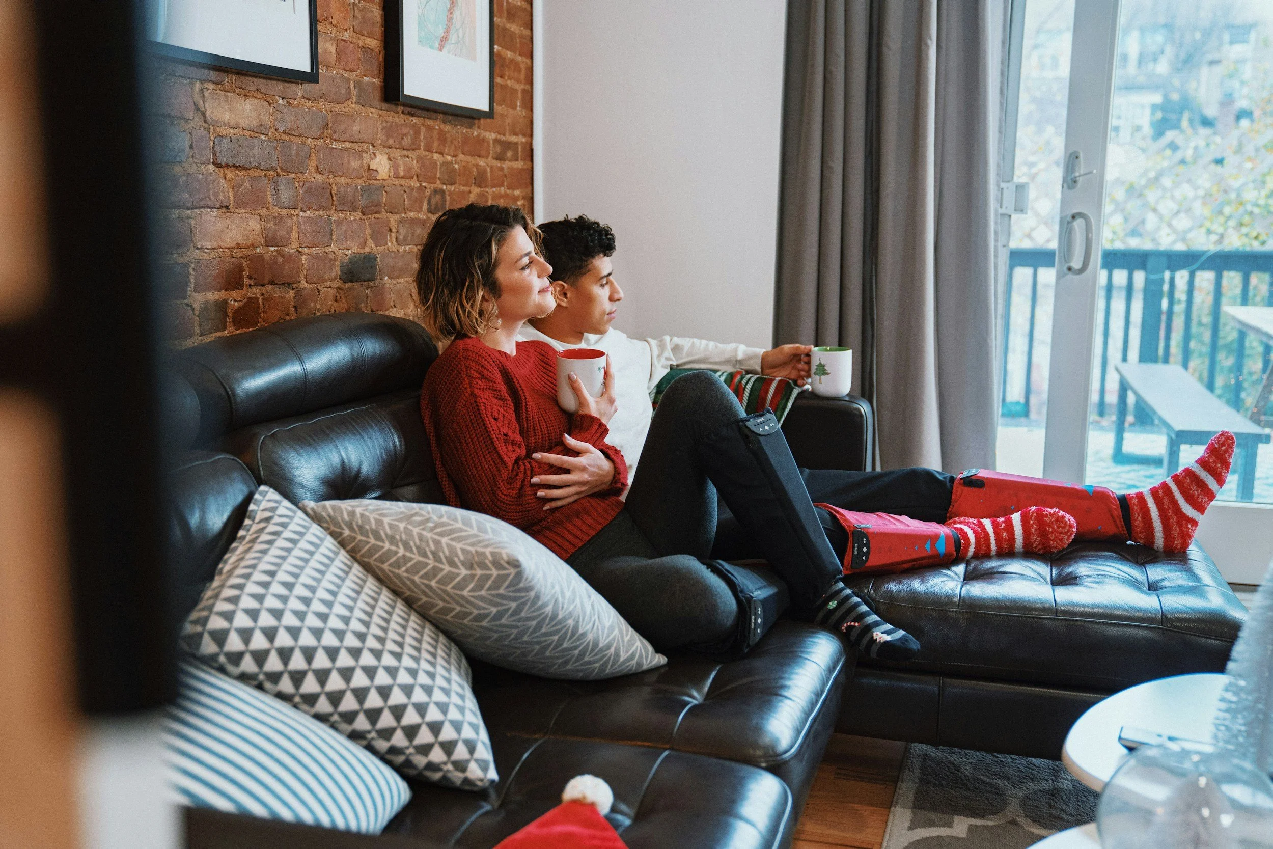 A woman and a boy sit on a black leather sofa, holding mugs, watching outside through glass sliding doors with a balcony visible. The woman is wearing a red sweater, and the boy is in white.