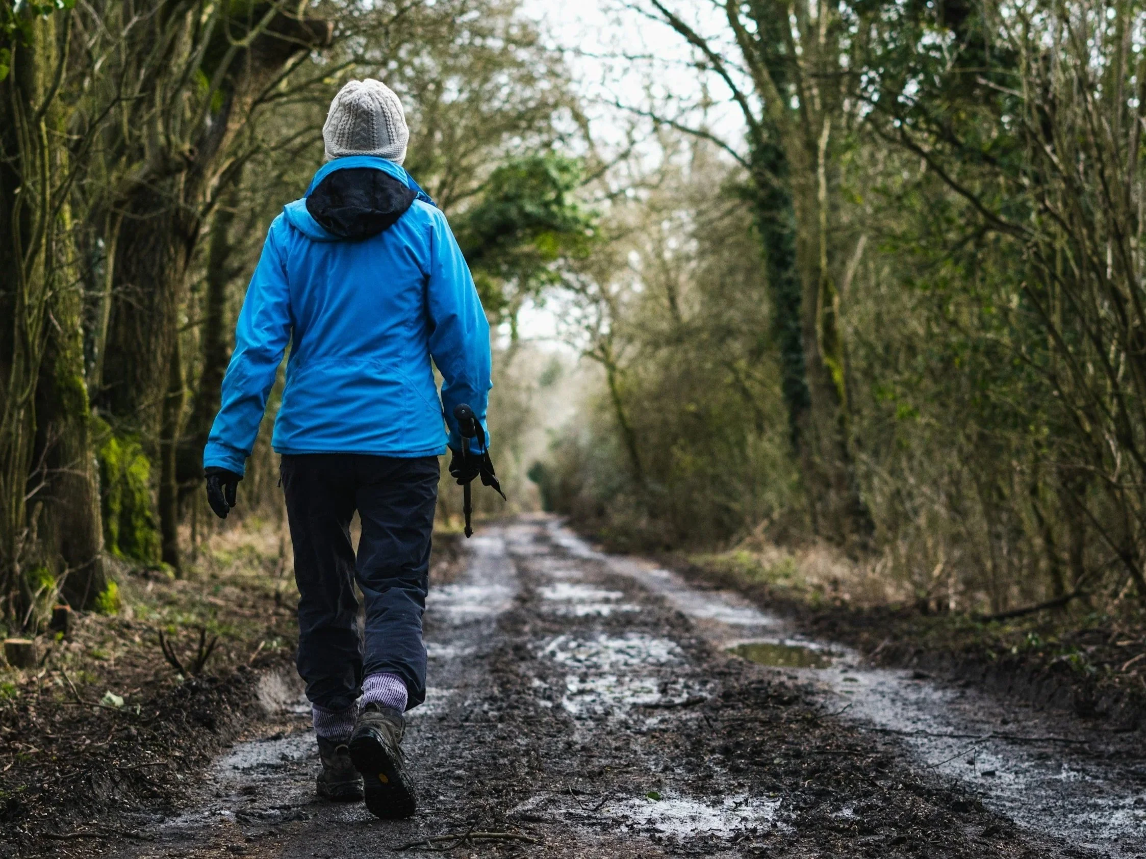 Person walking on a muddy forest trail wearing a blue jacket and gray beanie.