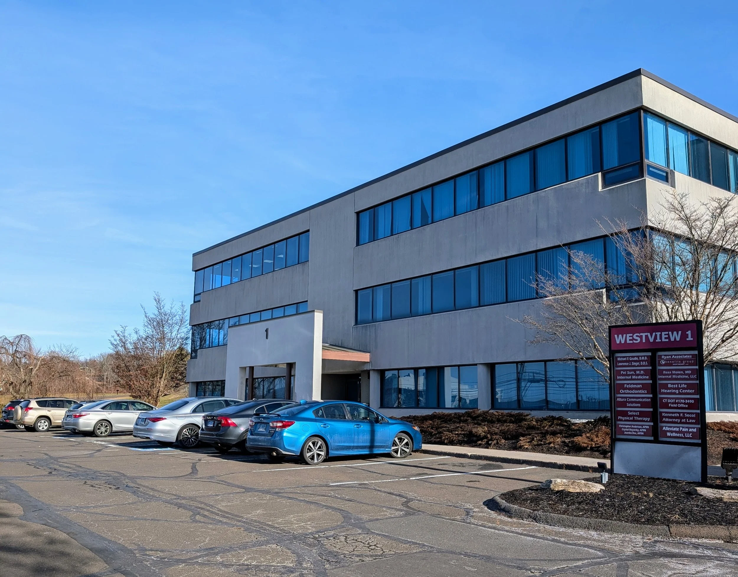 A three-story commercial office building with a parking lot in front, featuring several cars parked. The building has large blue-tinted windows and a white facade. There's a sign that reads 850 North Main Street Extension, Bldg-1, 2C Wallingford, CT
