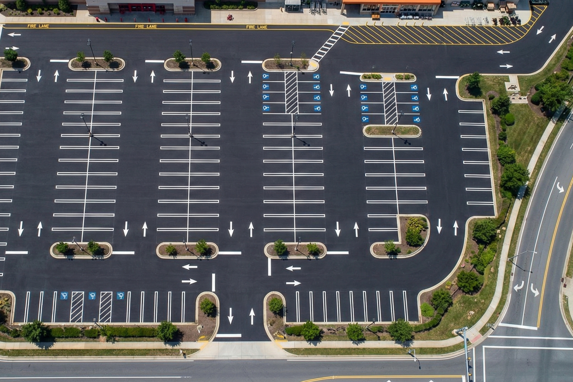 vue aérienne d'un parking presque vide avec des lignes blanches, des espaces pour handicapés, des îlots avec des arbres et des lampadaires, entouré de routes.