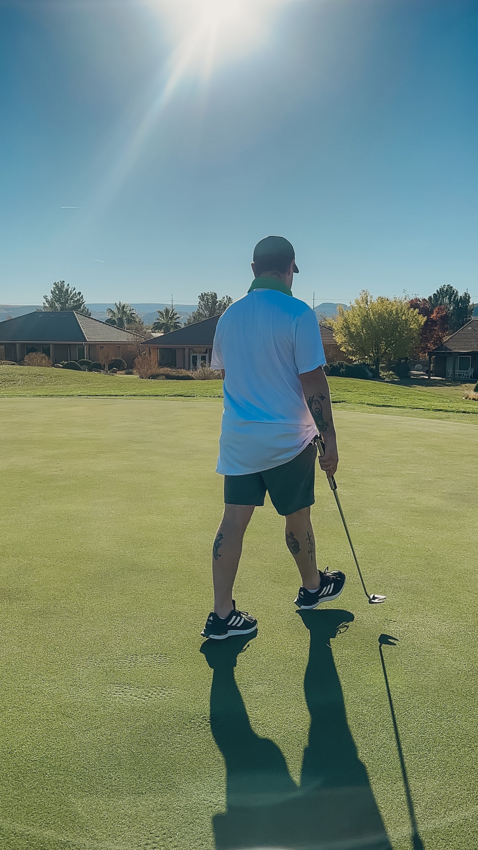 A man dressed in a white shirt, black shorts, and black Adidas sneakers stands on a golf course holding a putter. The sun is shining brightly overhead, casting a shadow of the man and his golf clubs on the green. Residential houses and trees are visible in the background.