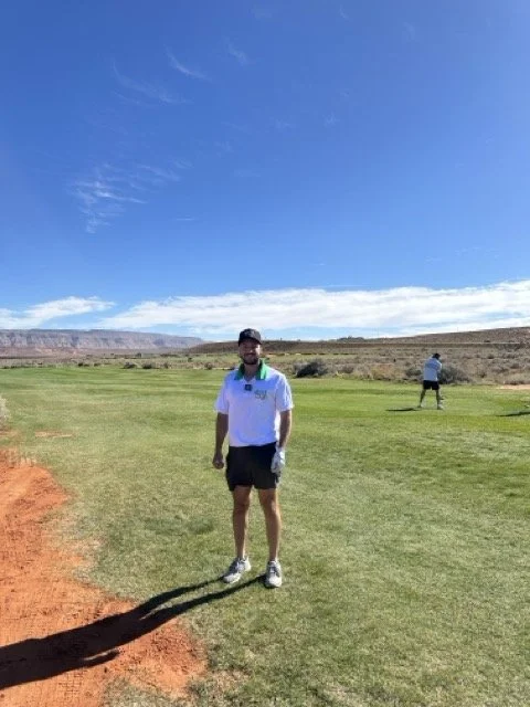 A man standing on a golf course with green grass, desert landscape, and blue sky with clouds in the background.