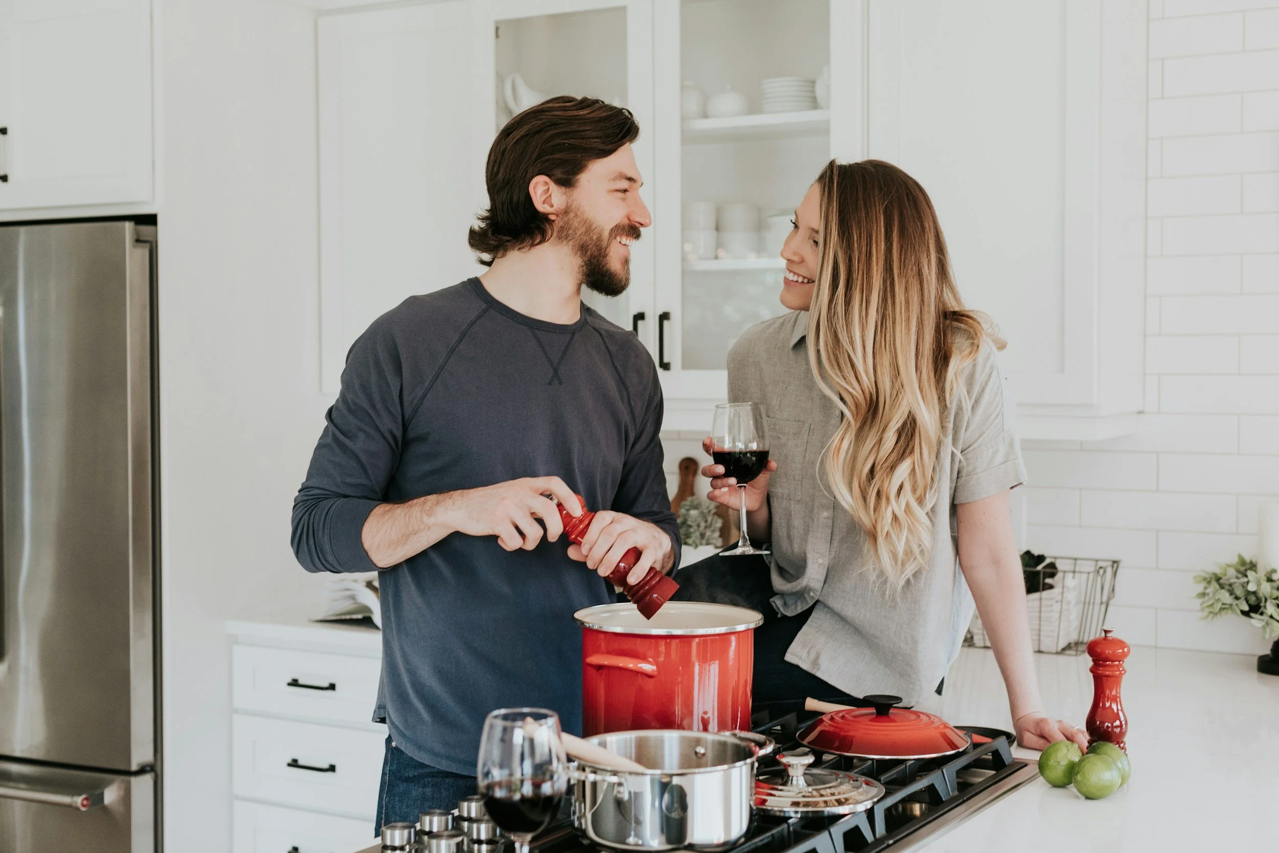 A couple cooking together in a modern kitchen, smiling at each other, with the woman holding a glass of red wine and the man adding seasoning to a pot.