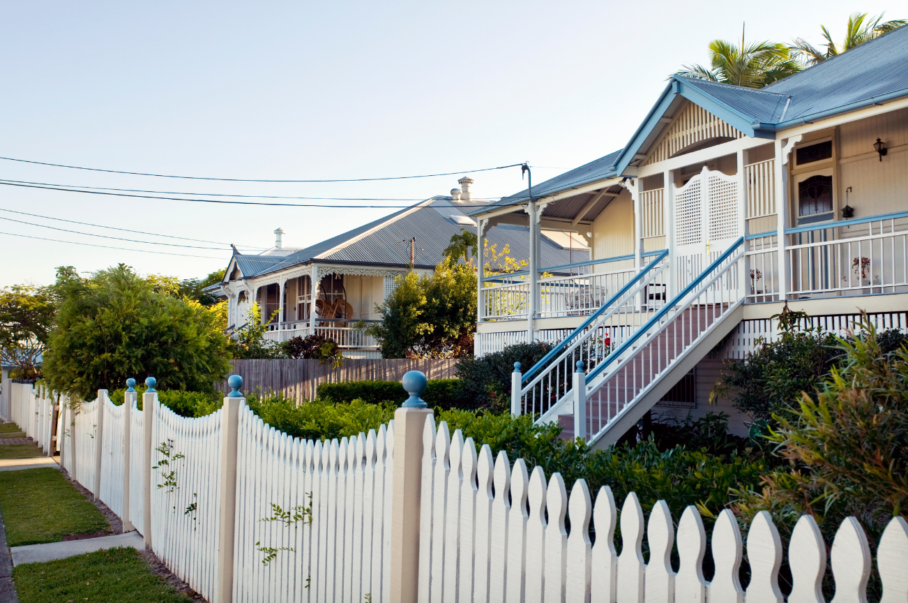 White Victorian style house with a staircase, surrounded by a white picket fence and lush greenery, under a clear sky.