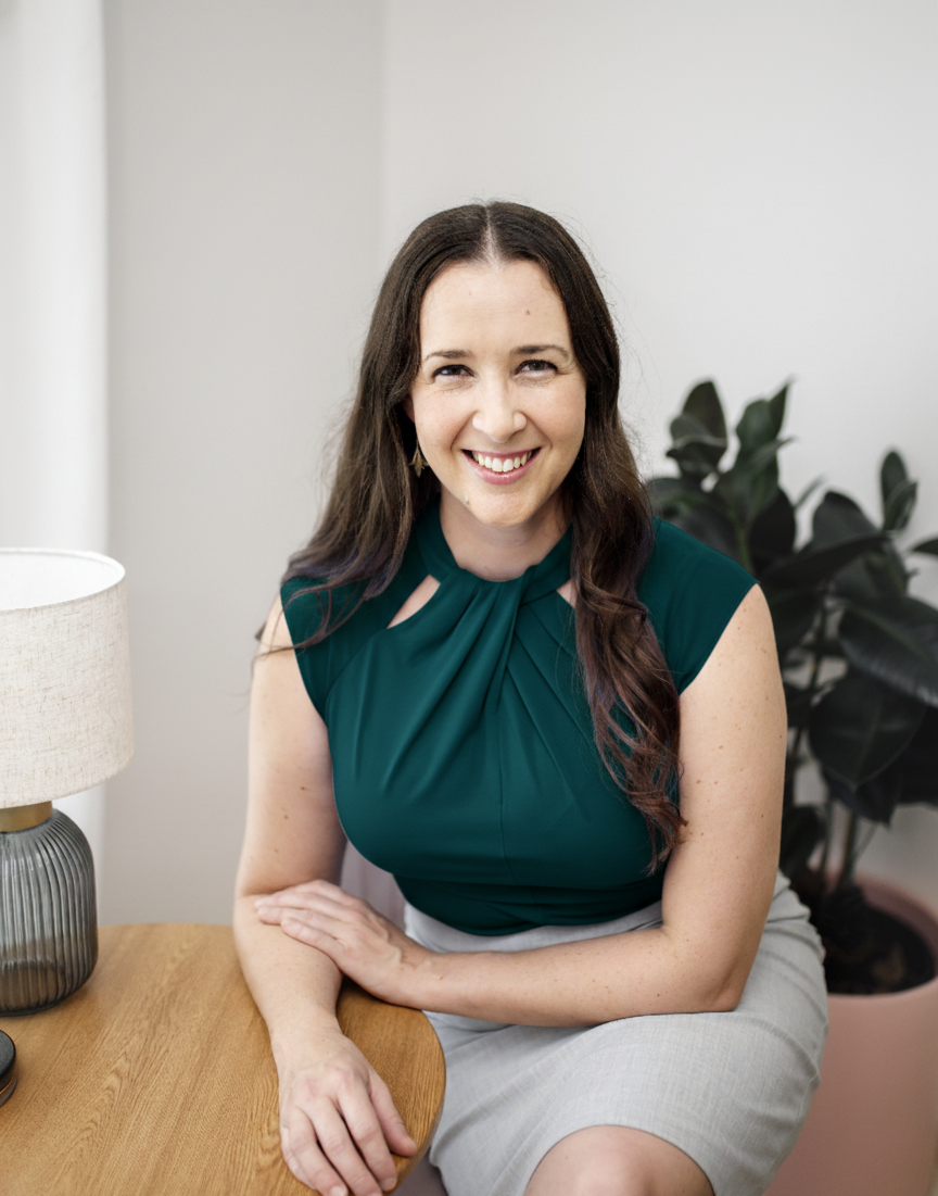 A smiling woman with long brown hair, wearing a dark green top and light gray skirt, sitting next to a wooden table and a lamp, with a plant in the background.