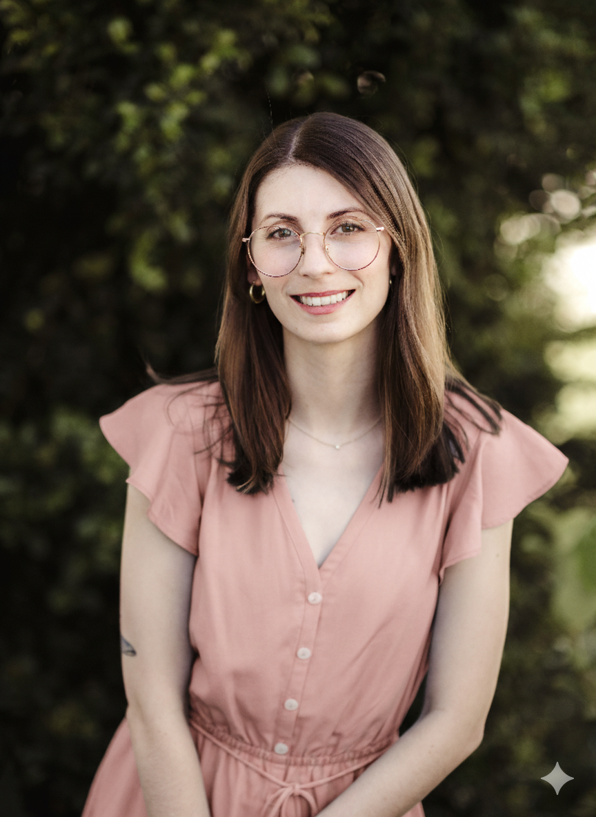 Young woman with shoulder-length brown hair, glasses, hoop earrings, and a delicate necklace, smiling outdoors in front of green foliage, wearing a pink button-down dress.
