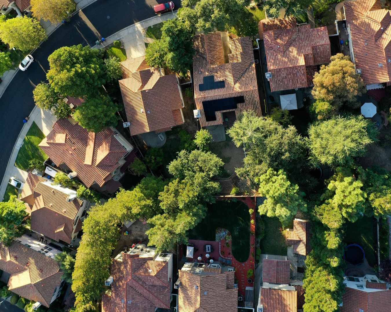 Aerial view of suburban neighborhood with terracotta-roofed houses nestled among lush green trees. Curved asphalt road and gardens add a serene vibe.
