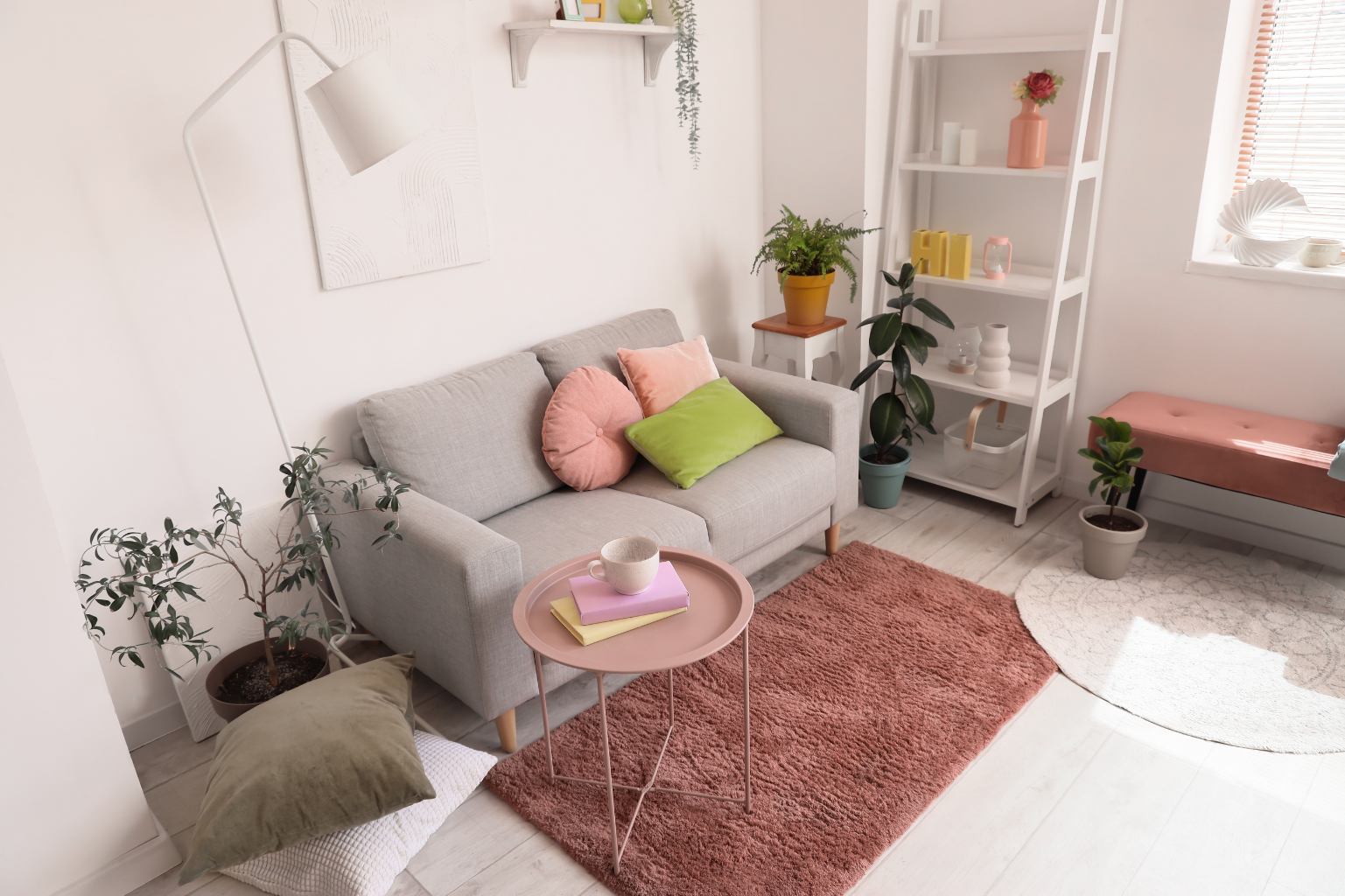 Bright living room with light-colored sofa, pink and green pillows, pink round side table with books and mug, potted plants, white shelving unit, pink bench, and window with blinds.