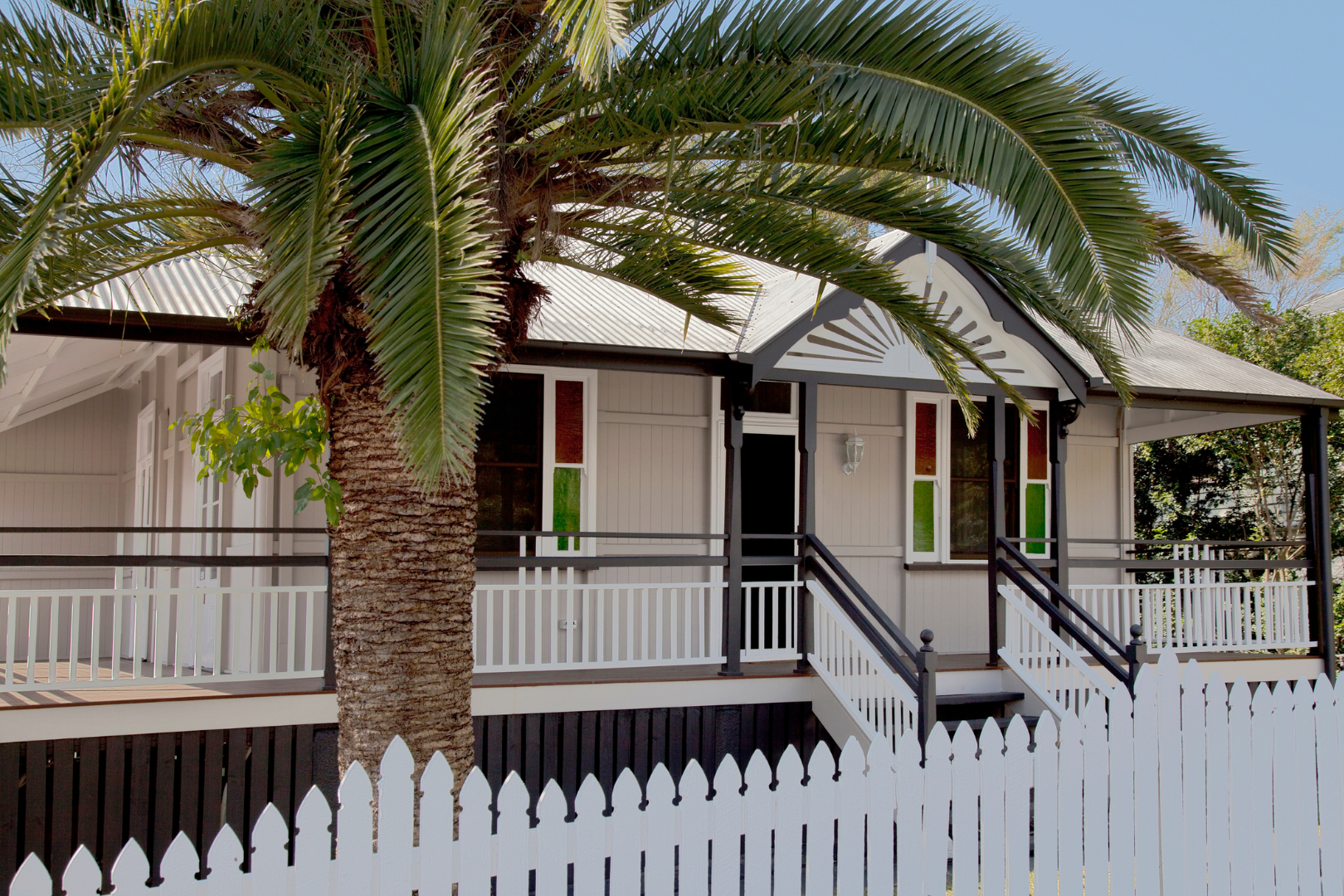 A house with a wrapping porch, black and white railing, a steep gabled roof, and stained glass windows. There's a large palm tree in the front yard and a white picket fence.