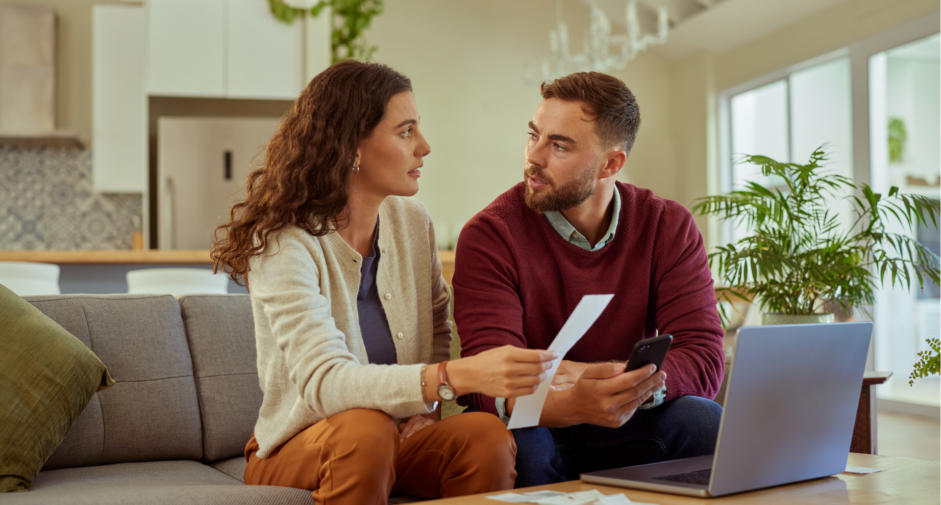 A man and woman sitting on a sofa in a well-lit living room, engaged in a serious conversation. The woman holds a piece of paper and the man holds a smartphone. There's a laptop and some papers on the table in front of them, with indoor plants and a kitchen in the background.