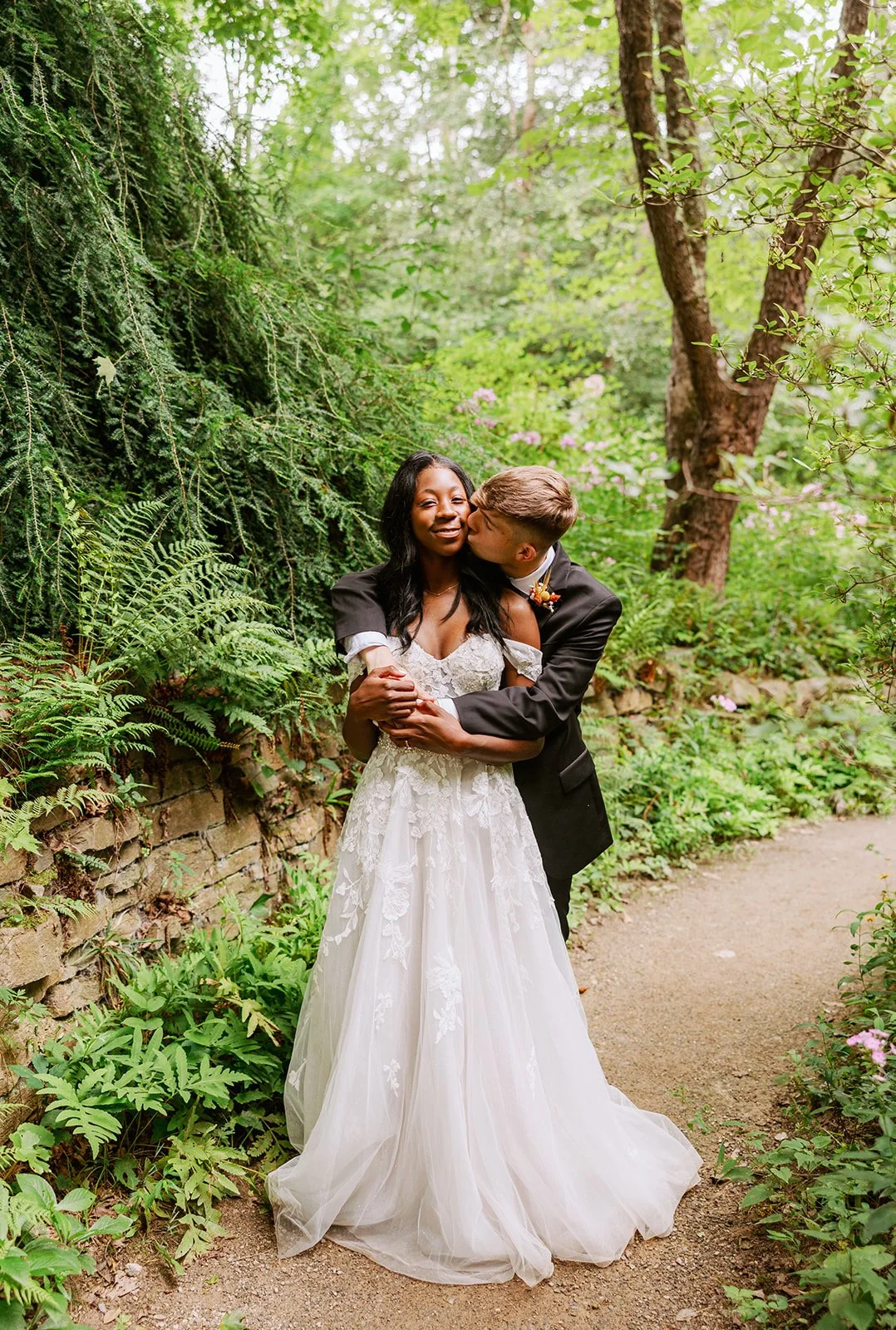 A newlywed couple stands on a garden path surrounded by green foliage. The bride wears a white lace wedding gown and smiles as the groom, in a black tuxedo, kisses her cheek. The scene is lush and romantic.