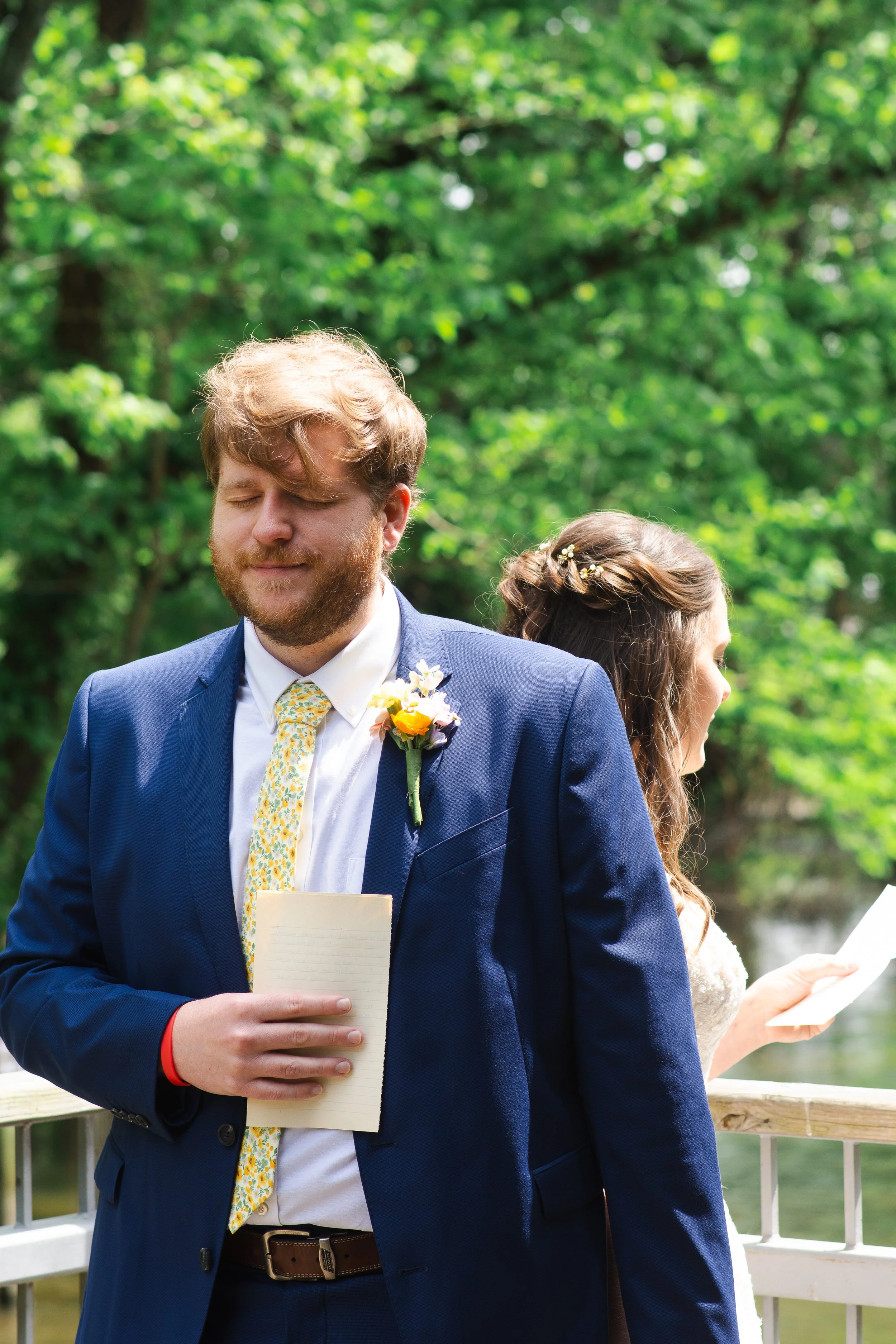 A man in a blue suit with a floral tie and a boutonniere, holding a notepad and standing outdoors next to a woman in a wedding dress, on a wooden deck with green trees in the background.