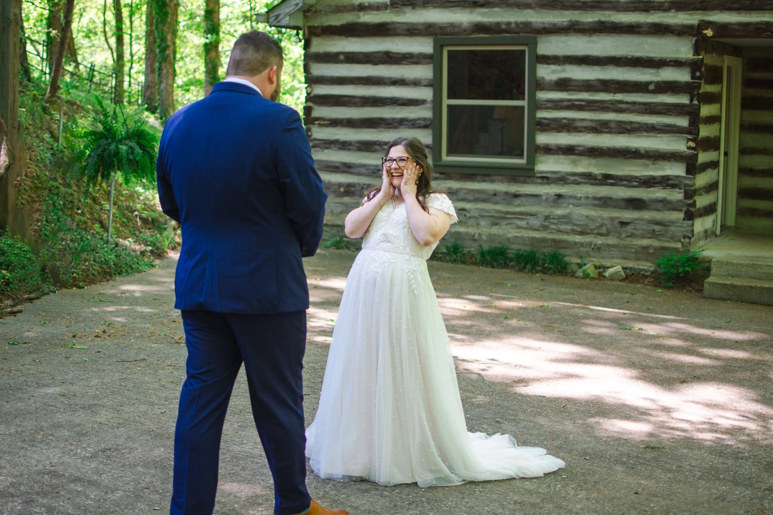 A bride in a white wedding dress and a groom in a blue suit standing outdoors near a log cabin in a wooded area. The bride is smiling with her hands on her cheeks, while the groom is facing her, wearing brown shoes.