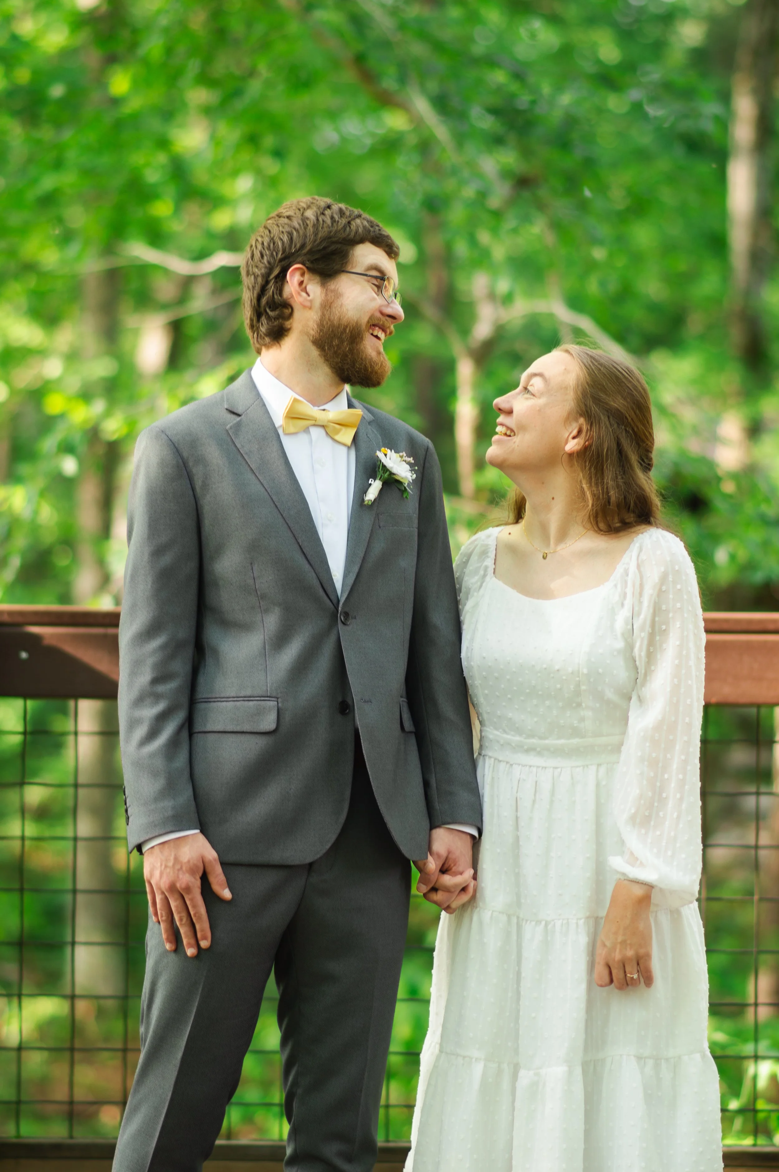 A couple holding hands and smiling at each other during their wedding outdoors surrounded by green trees.