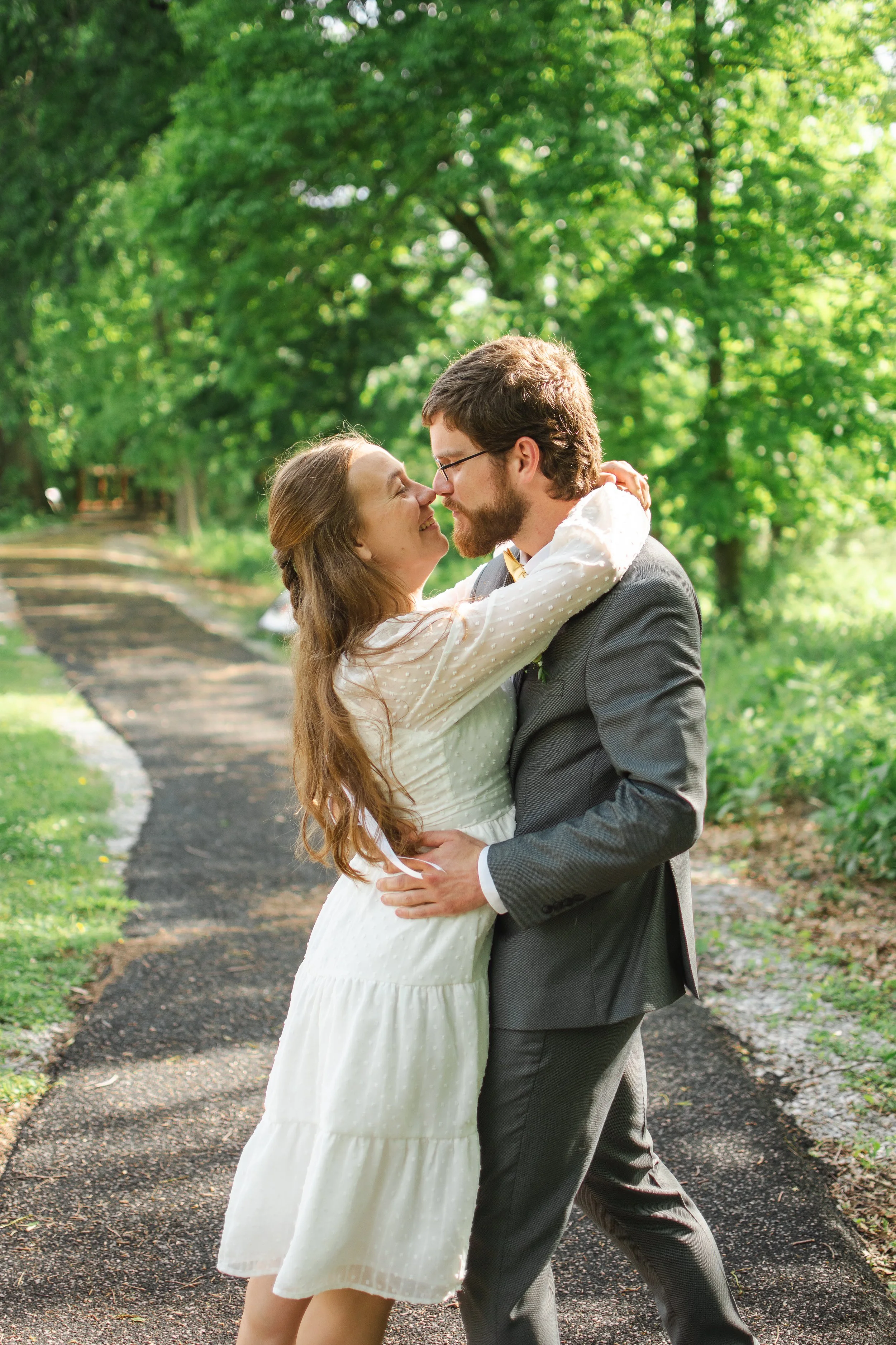A couple in wedding attire embracing each other on a forest path with lush green trees in the background.