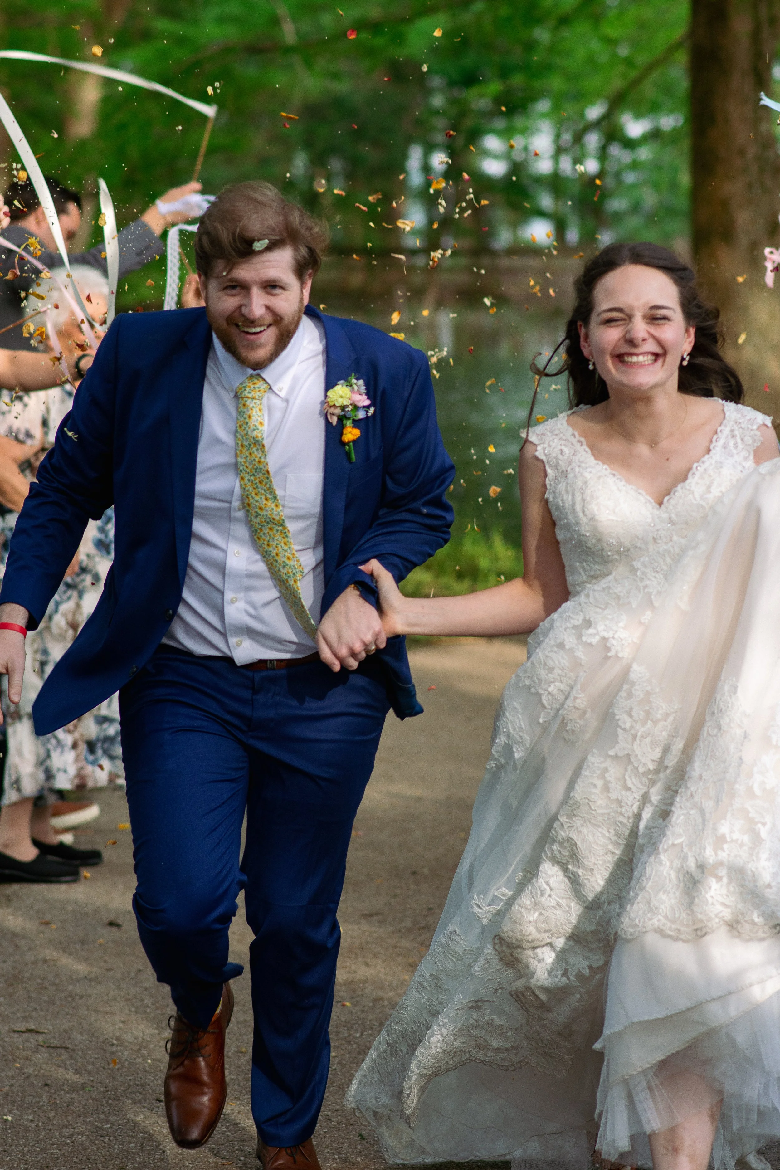 A newlywed couple, holding hands and smiling, running outdoors with wedding guests throwing confetti in the background.