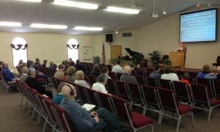 People attending a church service or presentation in a room with chairs, with a piano and a screen displaying a presentation.