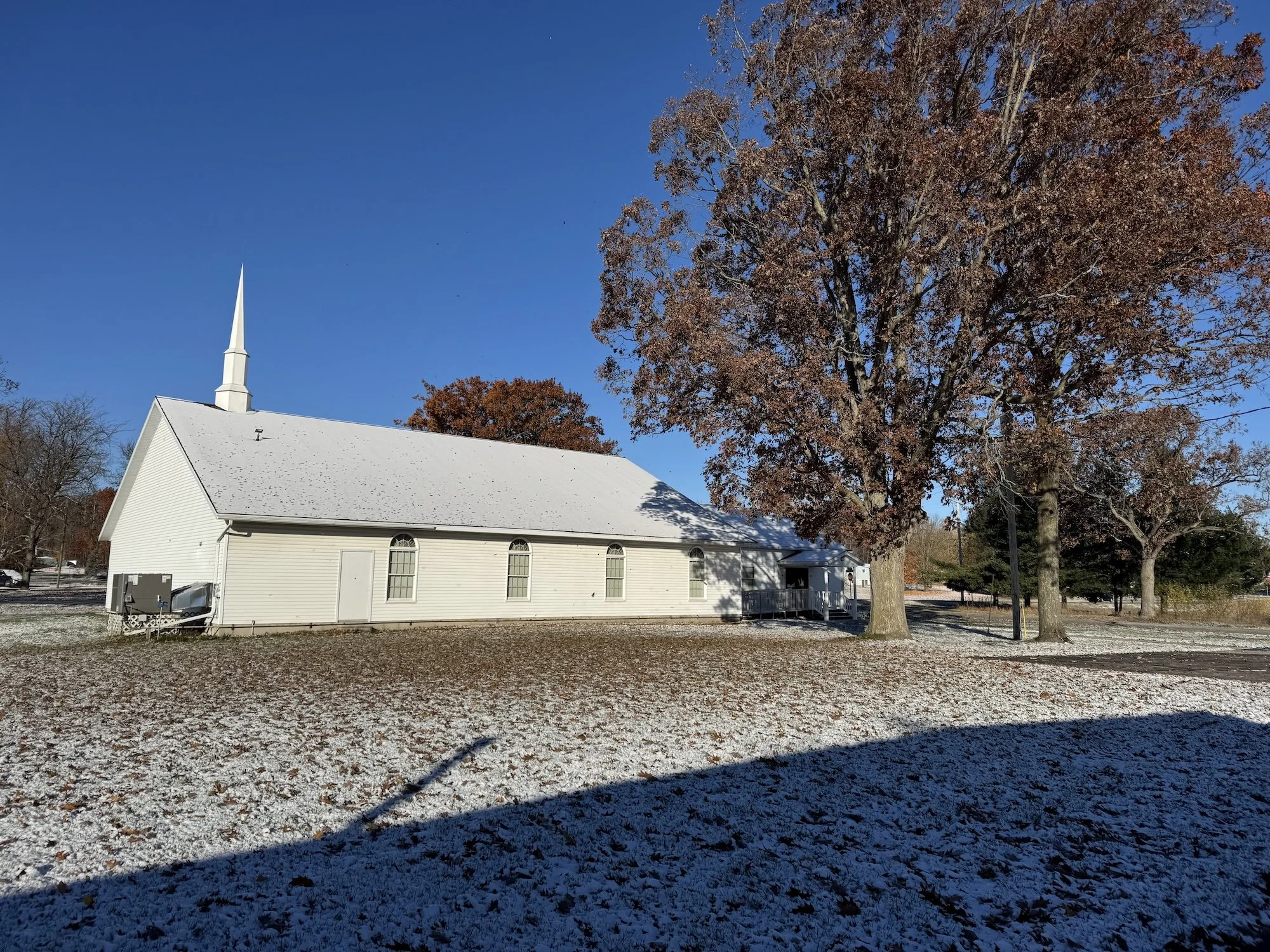 White church with a steeple, surrounded by leafless and brown trees, under a clear blue sky, with a light dusting of snow on the ground.
