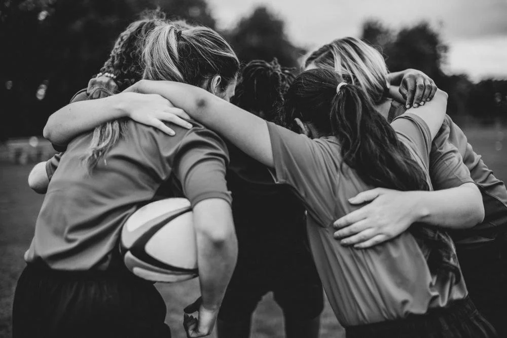 A group of female rugby players in a huddle before a game, with their arms around each other's shoulders.