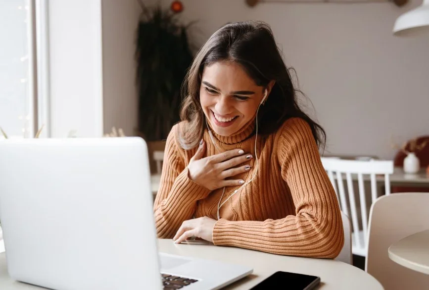 A woman smiling while sitting at a table with a laptop, using earphones, and touching her chest.