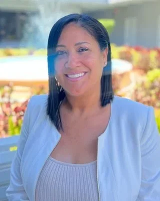 A woman with shoulder-length black hair smiling outdoors with flowers and a building in the background.